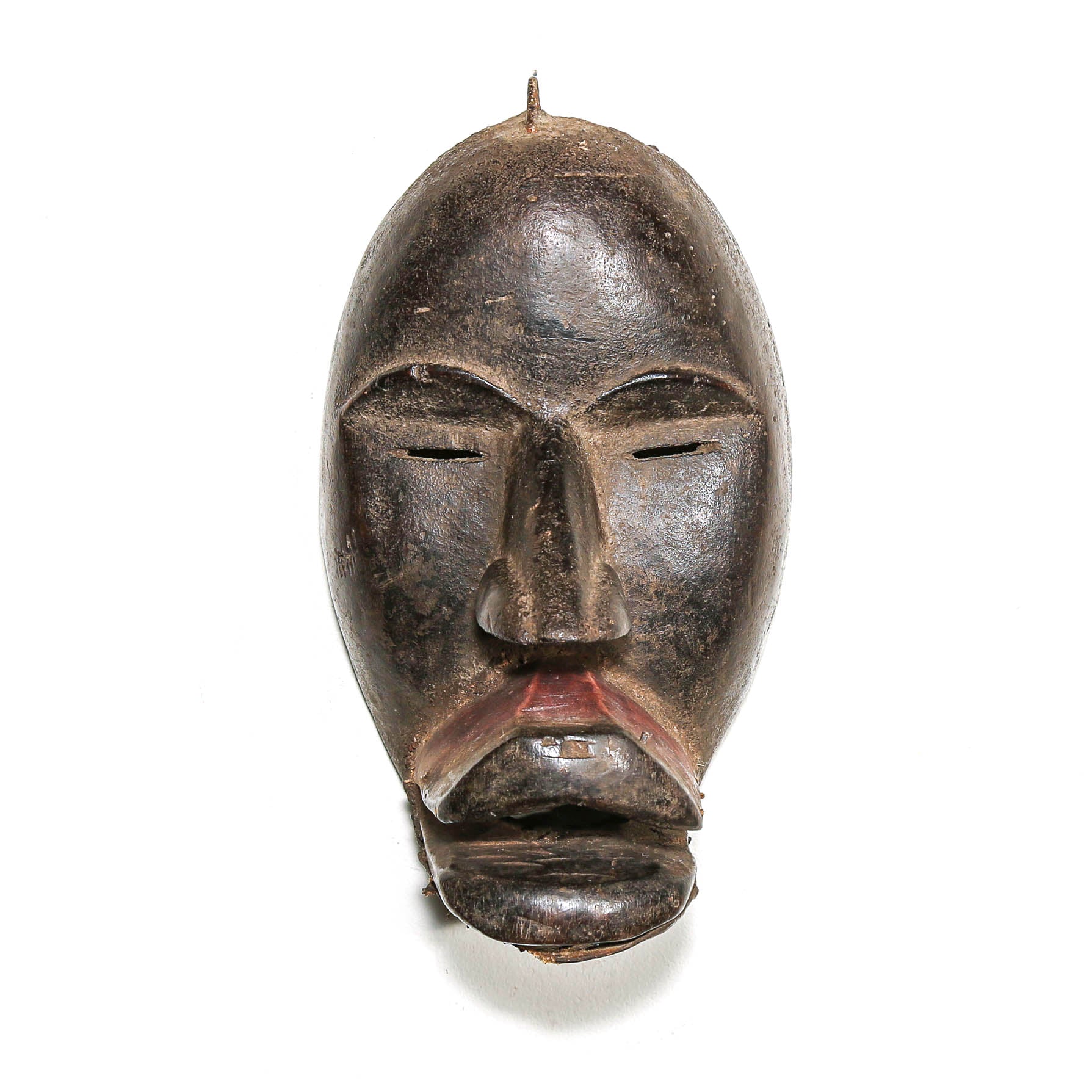 A dark carved wooden mask with a stylized face, closed eyes, and a small pointed top detail, photographed against a plain white background.
