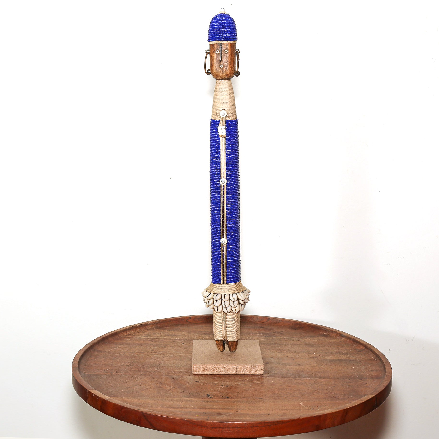 A hand-carved wooden African-style decorative figurine with a blue beaded body, displayed on a round wooden stand against a white background.