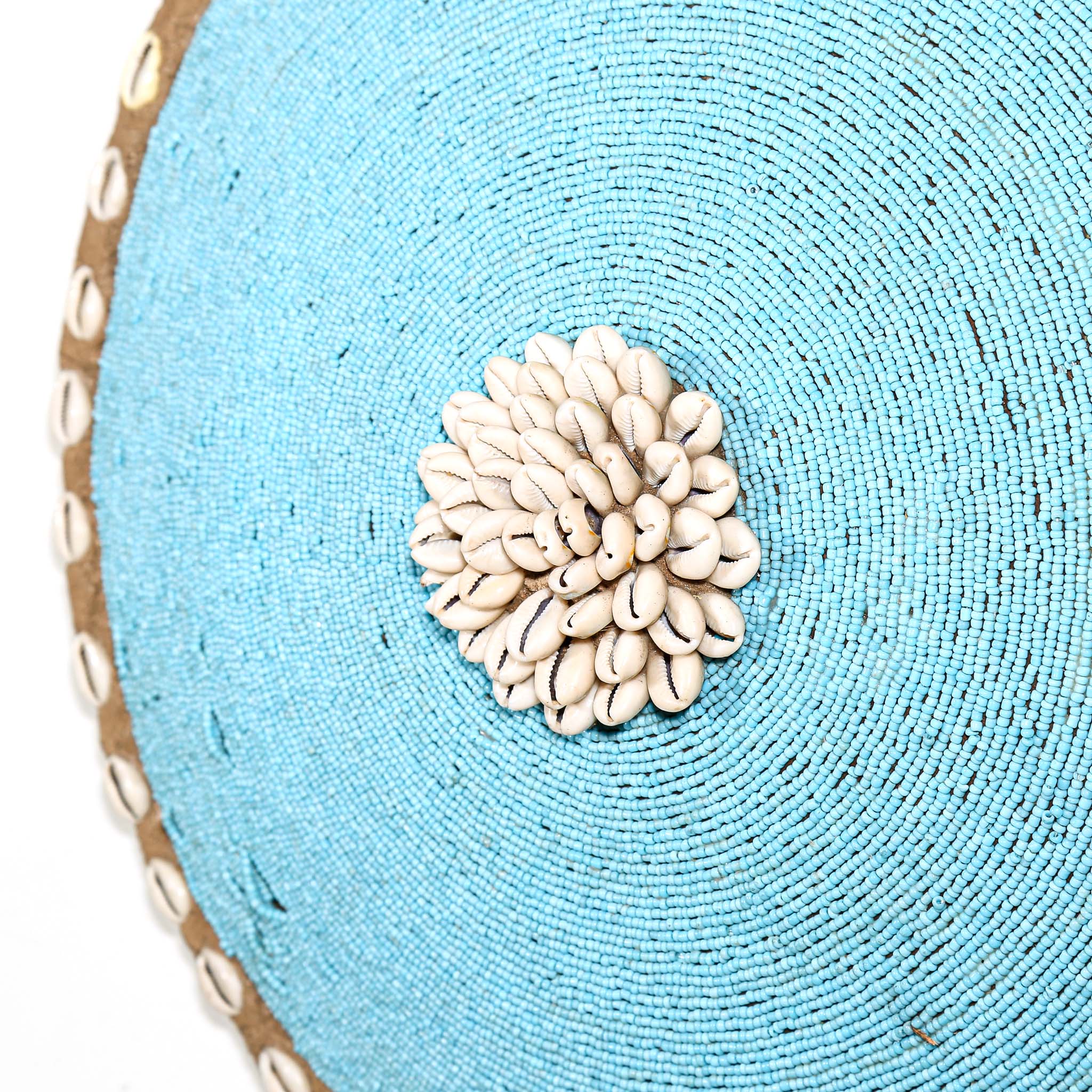 Round white beaded wall shield decor with a central white flower design made from cowrie shells hanging on a white wall.