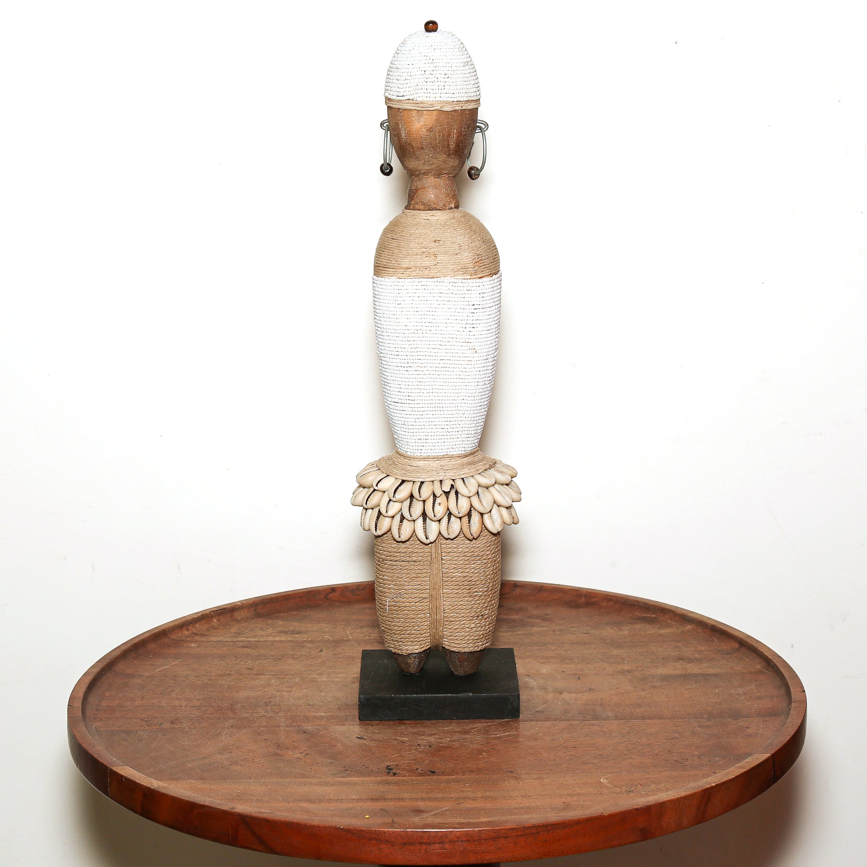 A handmade African wooden and woven doll sculpture displayed on a round wooden pedestal against a plain white background.