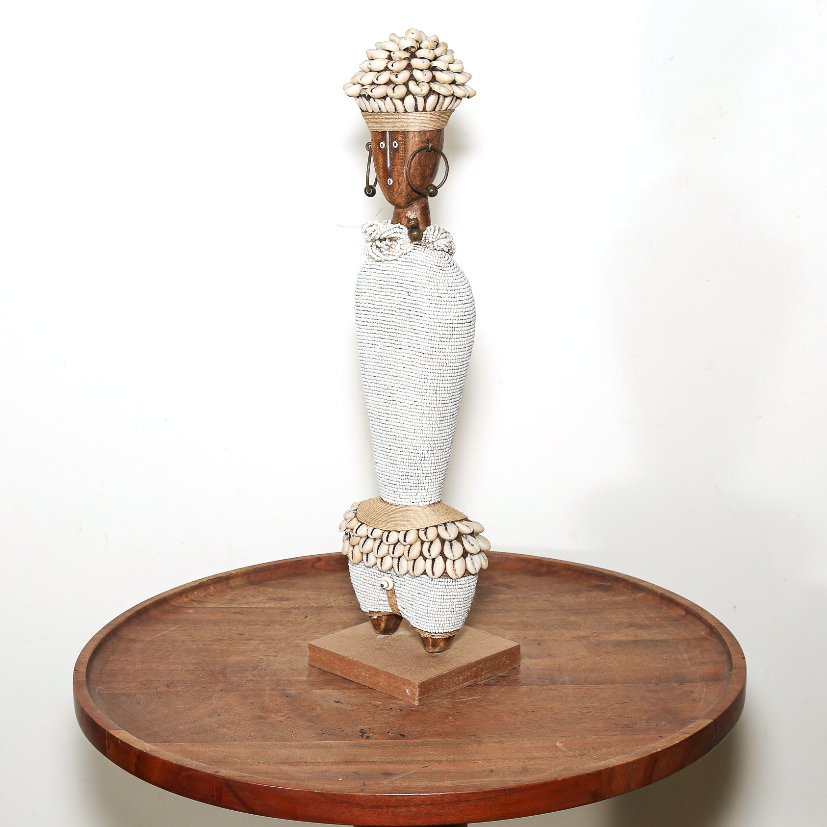A tall decorative sculpture of a stylized female figure made from white woven material, topped with a brown head and a cluster of small beige shells, displayed on a round wooden pedestal against a plain white background.