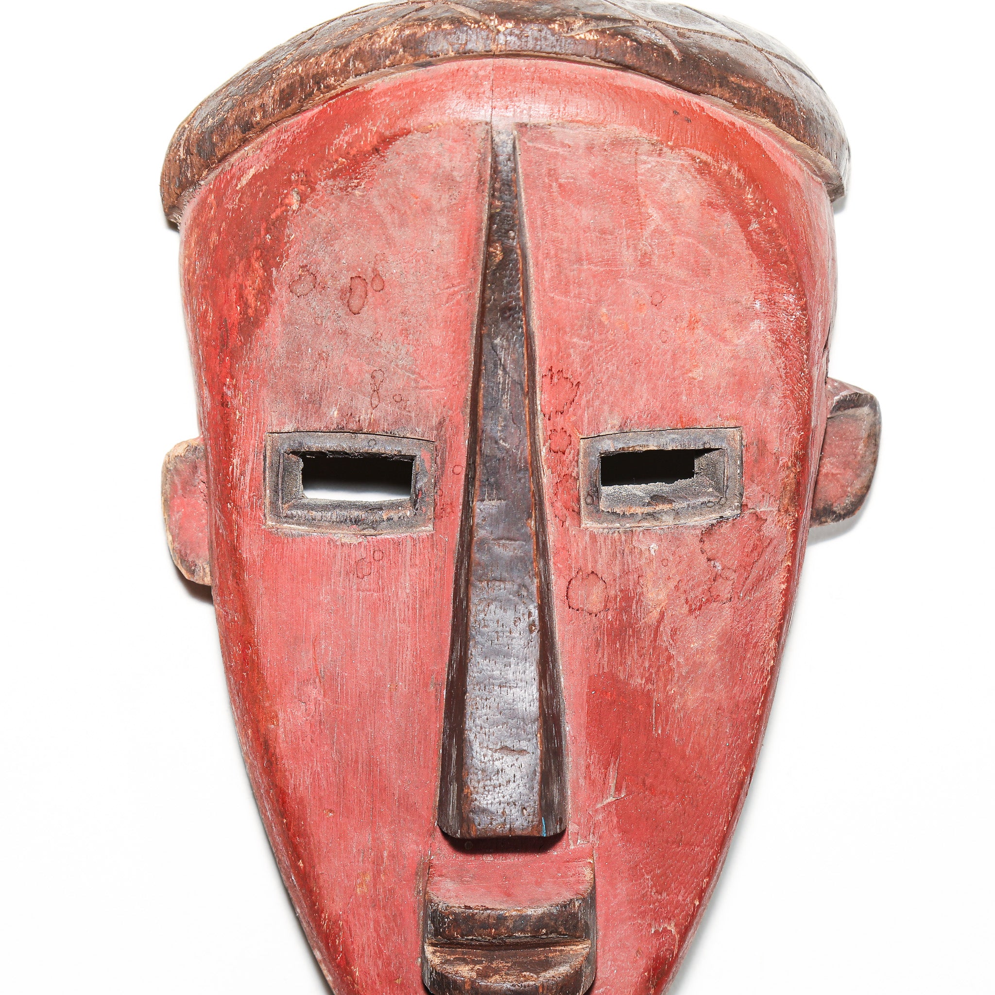 A traditional African wooden mask with a red-painted face, featuring a prominent dark wooden nose, rectangular eye slits, and a textured dark wooden top, shown against a plain white background.