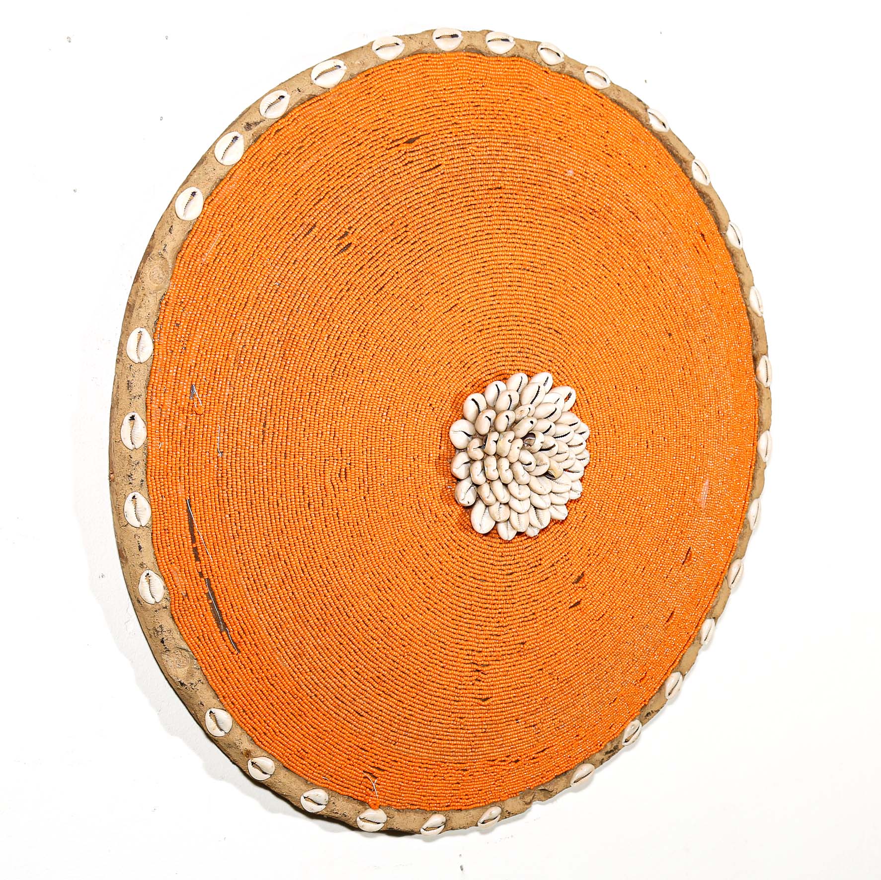 Round orange beaded wall shield decor with a central white flower design made from cowrie shells hanging on a white wall.