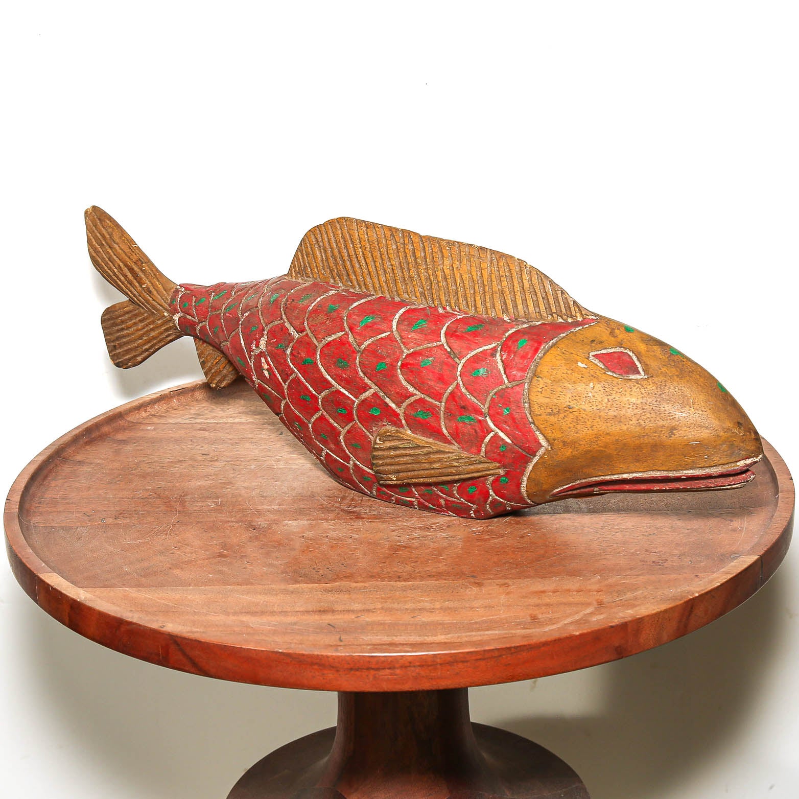 A hand-carved wooden fish sculpture with a red scaled pattern and natural wood head, displayed on a round wooden pedestal against a white background.