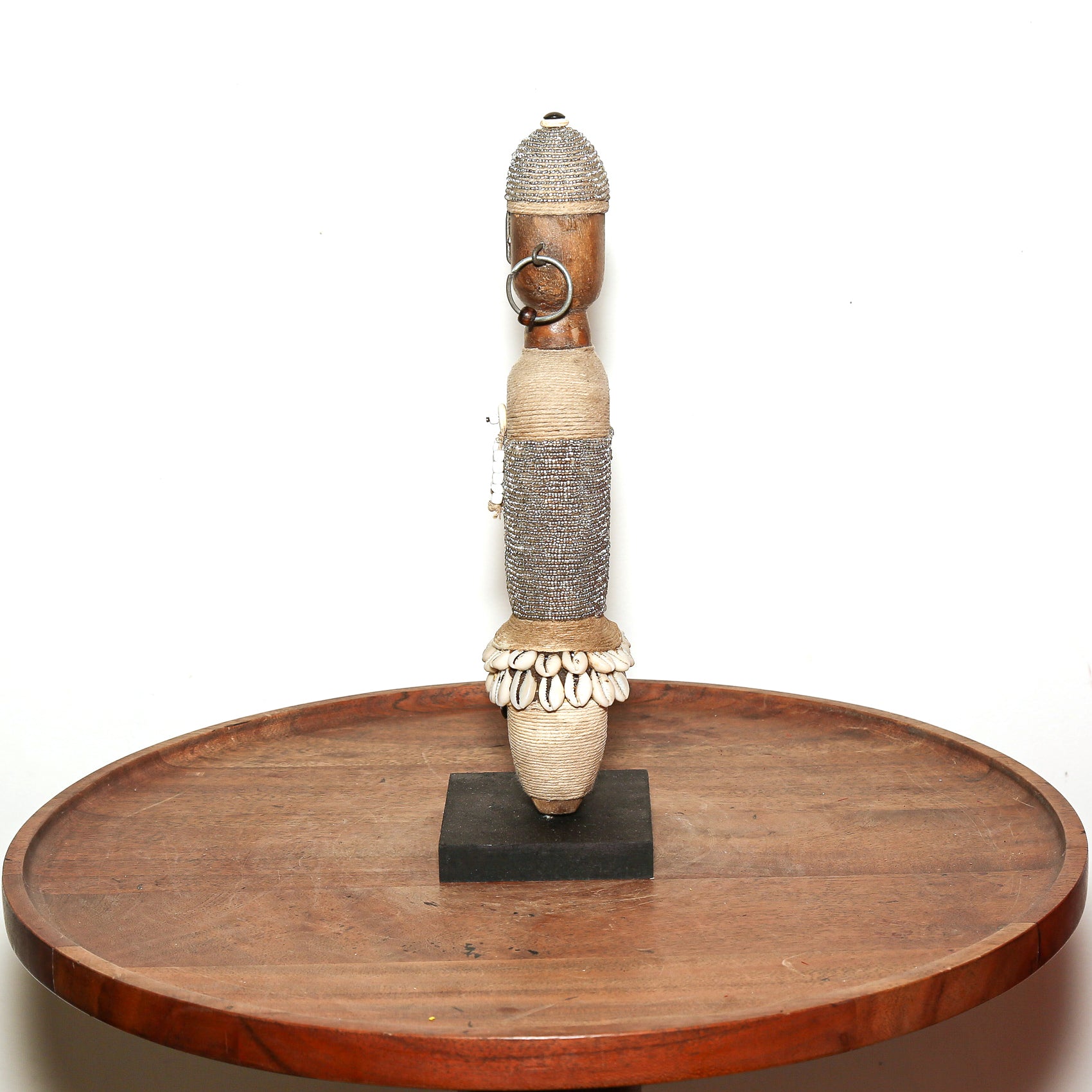 A tall, handcrafted African wooden sculpture with woven and shell details, displayed on a black base atop a round wooden table against a white background.