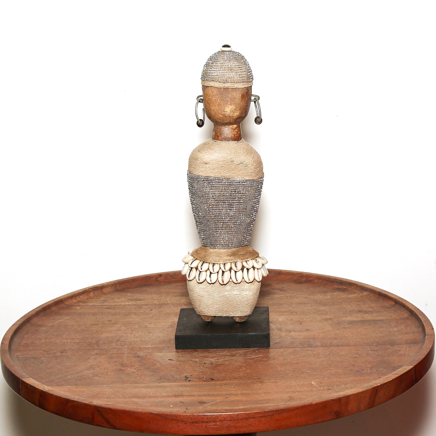 A traditional African wooden figurine with intricate beadwork and shell detailing, displayed on a round wooden table against a white background.