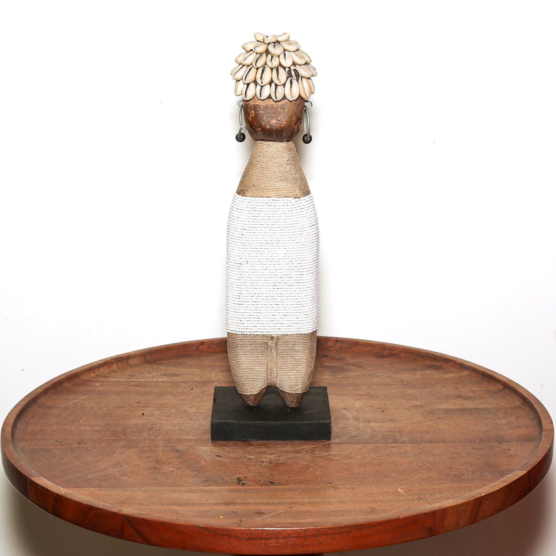 A handmade African-style wooden doll figurine with a woven white skirt, natural wood body, and a head decorated with cowrie shells, displayed on a round wooden tray against a white background.