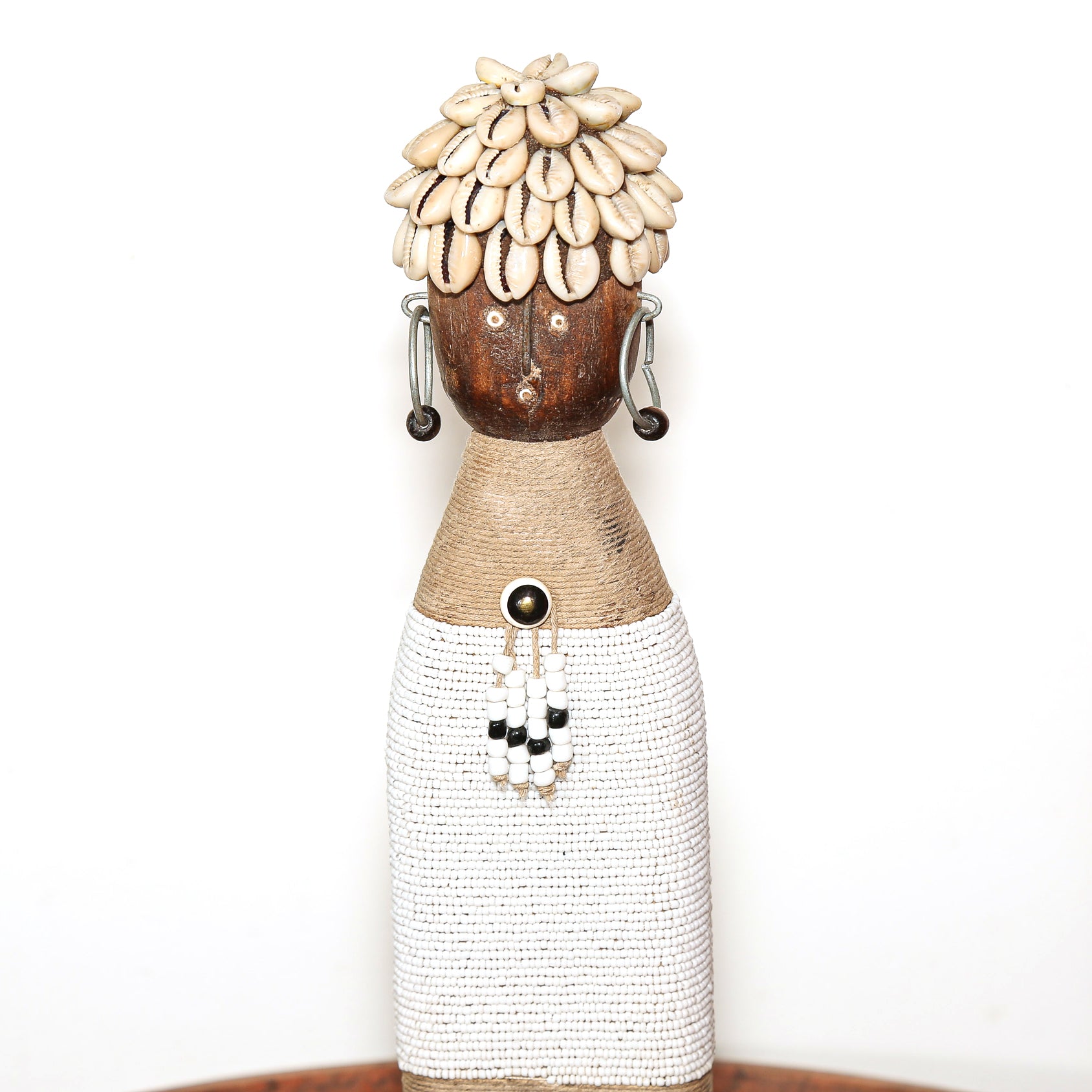 A decorative African-style doll figurine with a woven brown body, white beaded skirt, shell-like hair, and dangling earrings, photographed against a plain white background.