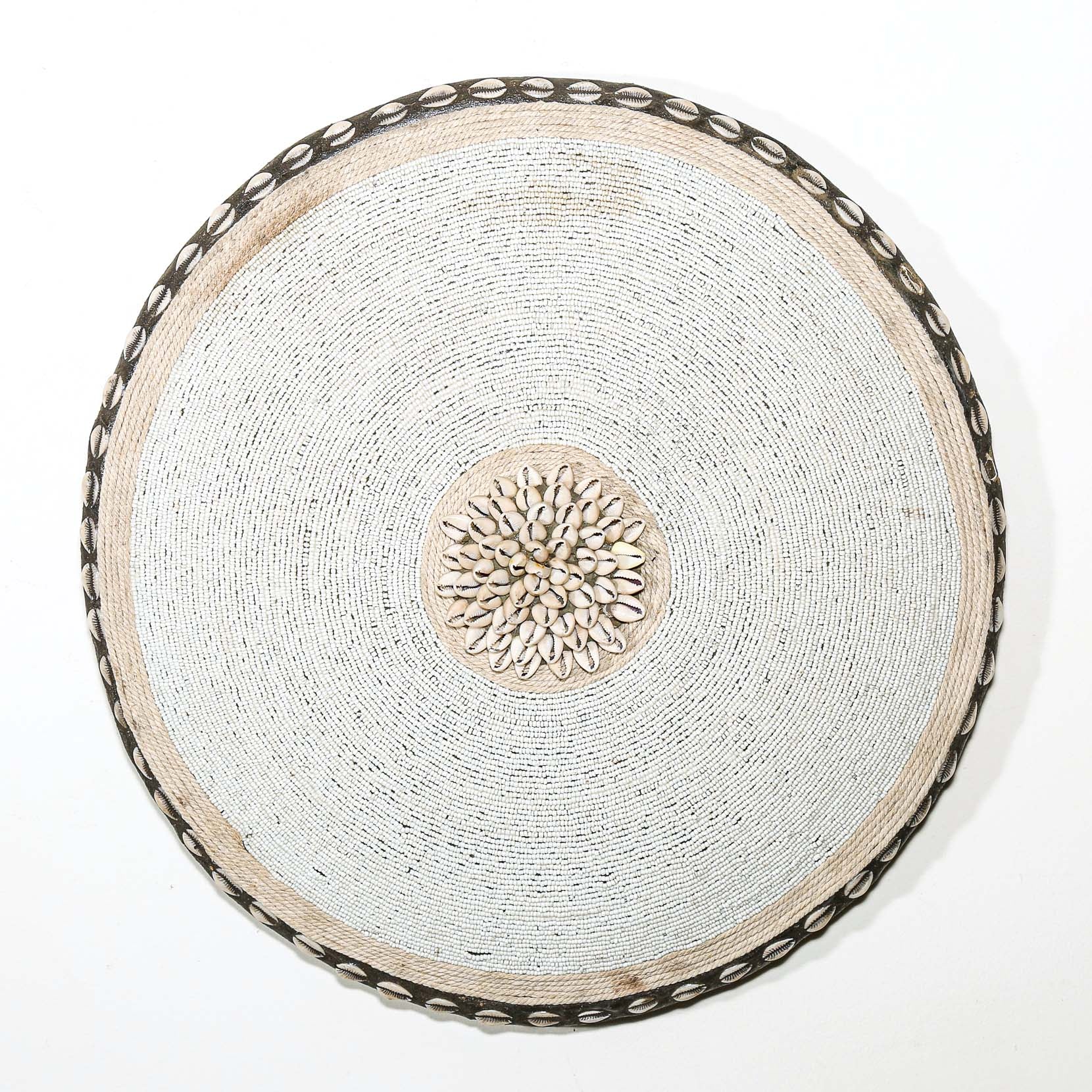 Round white beaded wall shield decor with a central white flower design made from cowrie shells hanging on a white wall.