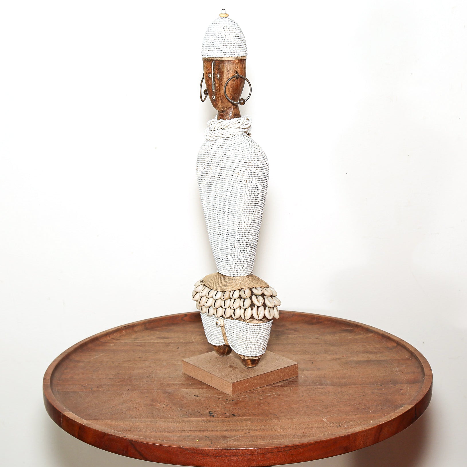 A tall African-style woven sculpture with a white textured body, brown wooden neck and feet, and circular metal earrings, displayed on a round wooden pedestal against a plain white background.