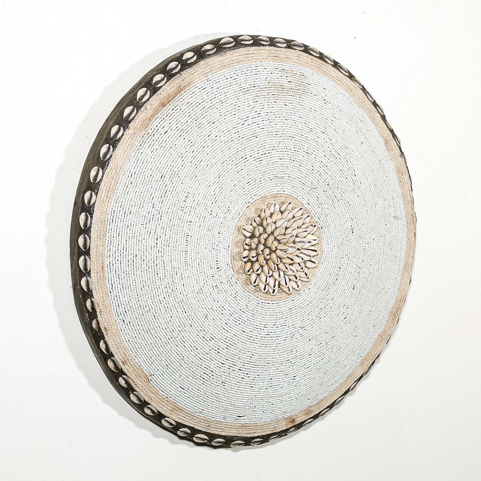 Round white beaded wall shield decor with a central white flower design made from cowrie shells hanging on a white wall.