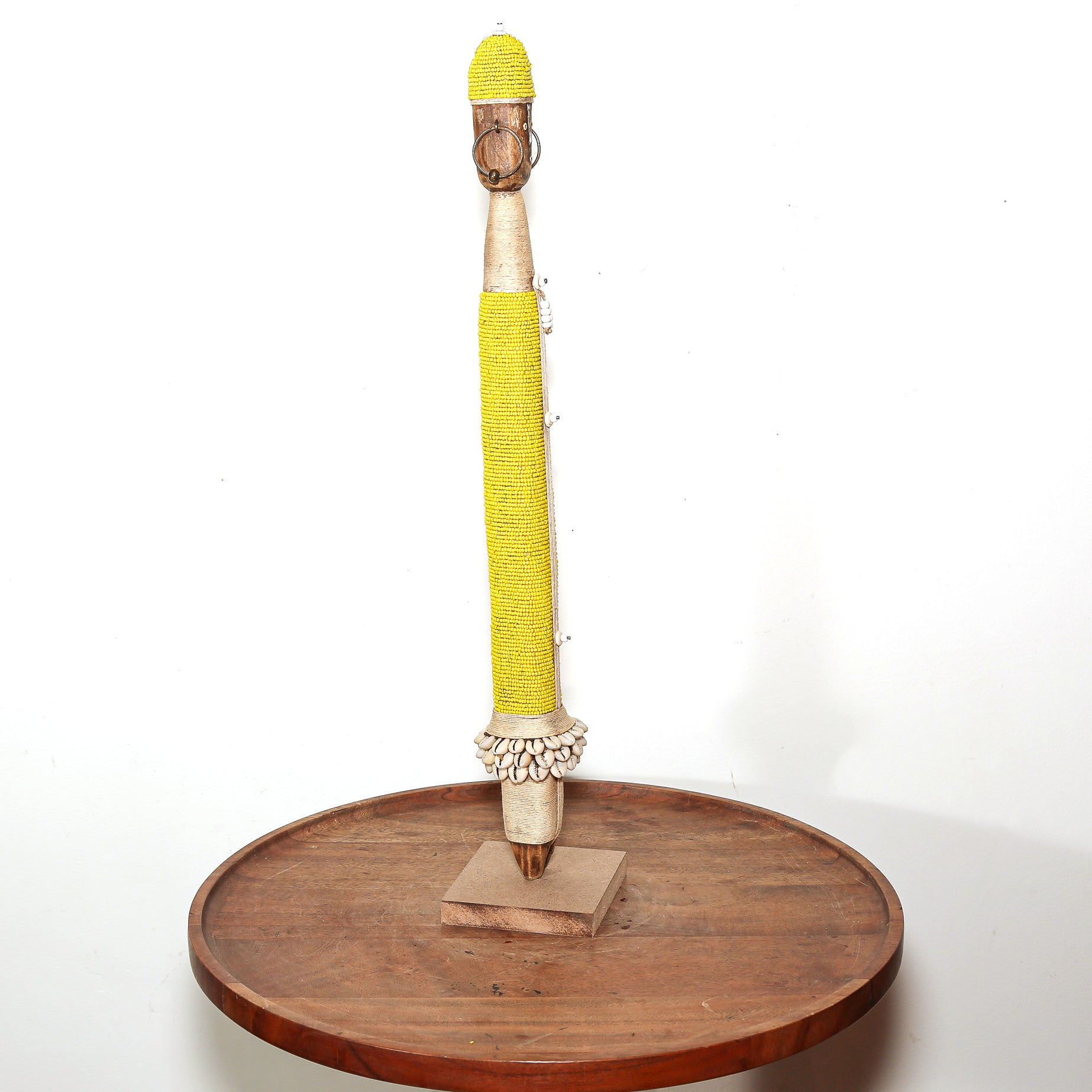 A decorative vertical object wrapped in bright yellow material, mounted on a small wooden base and set on a round wooden tray against a plain white background.