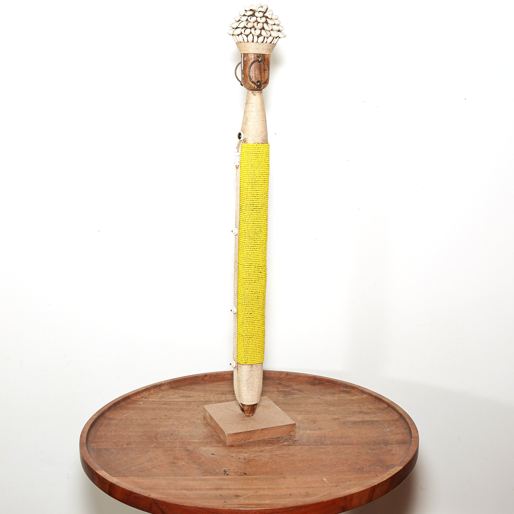 A handmade wooden staff or pole wrapped with bright yellow fabric in the center, featuring a decorative cluster of small white beads or shells at the top, displayed on a round wooden stand against a plain white background.