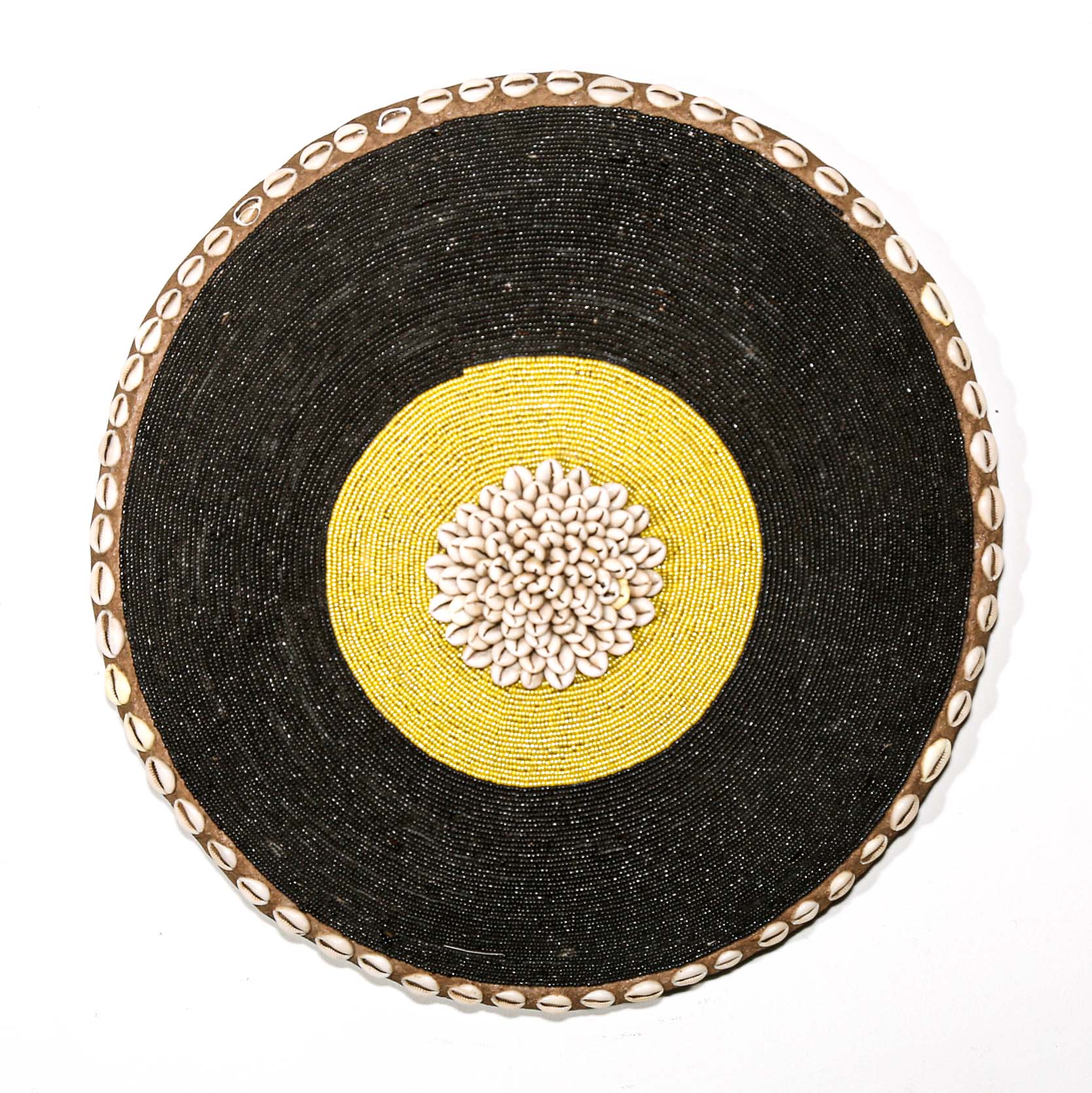 Round woven beaded shield with black surface, yellow center, and decorative edge hanging on a white wall
