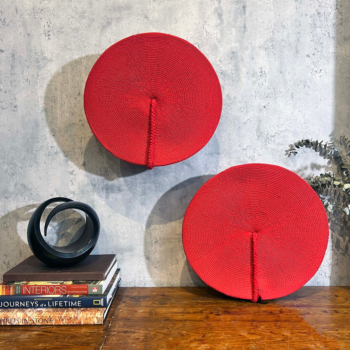 Two round red woven wall art pieces displayed on a rustic wooden table against a textured gray concrete wall, styled with books and a black decorative sculpture.