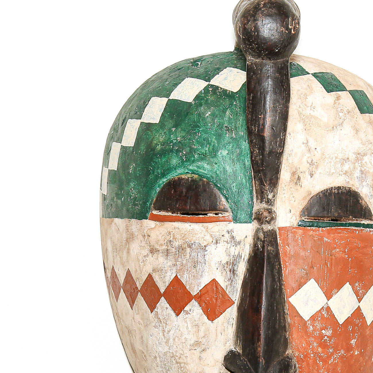 A close-up of a hand-painted African wooden mask with a green and white checkered pattern on one half, an orange diamond pattern on the lower half, and a dark central vertical feature, set against a plain white background.