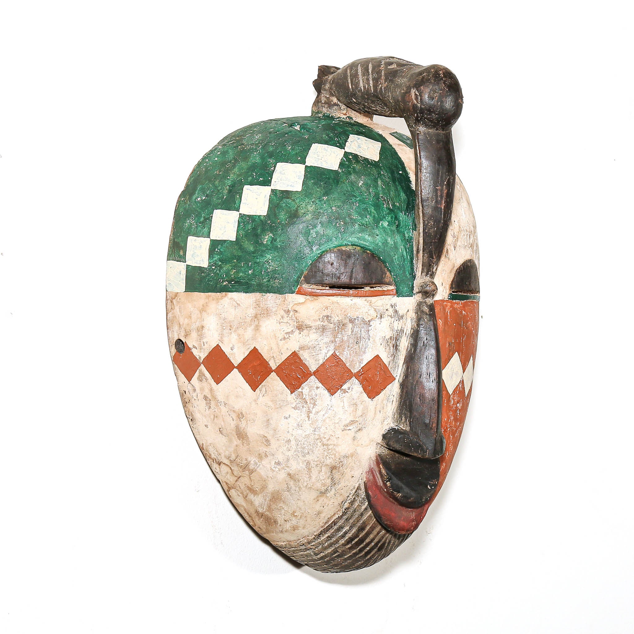 A hand-carved African wooden mask with a green and white diamond pattern on the forehead, a red diamond pattern on the cheek, and a dark brown handle on top, shown against a plain white background.