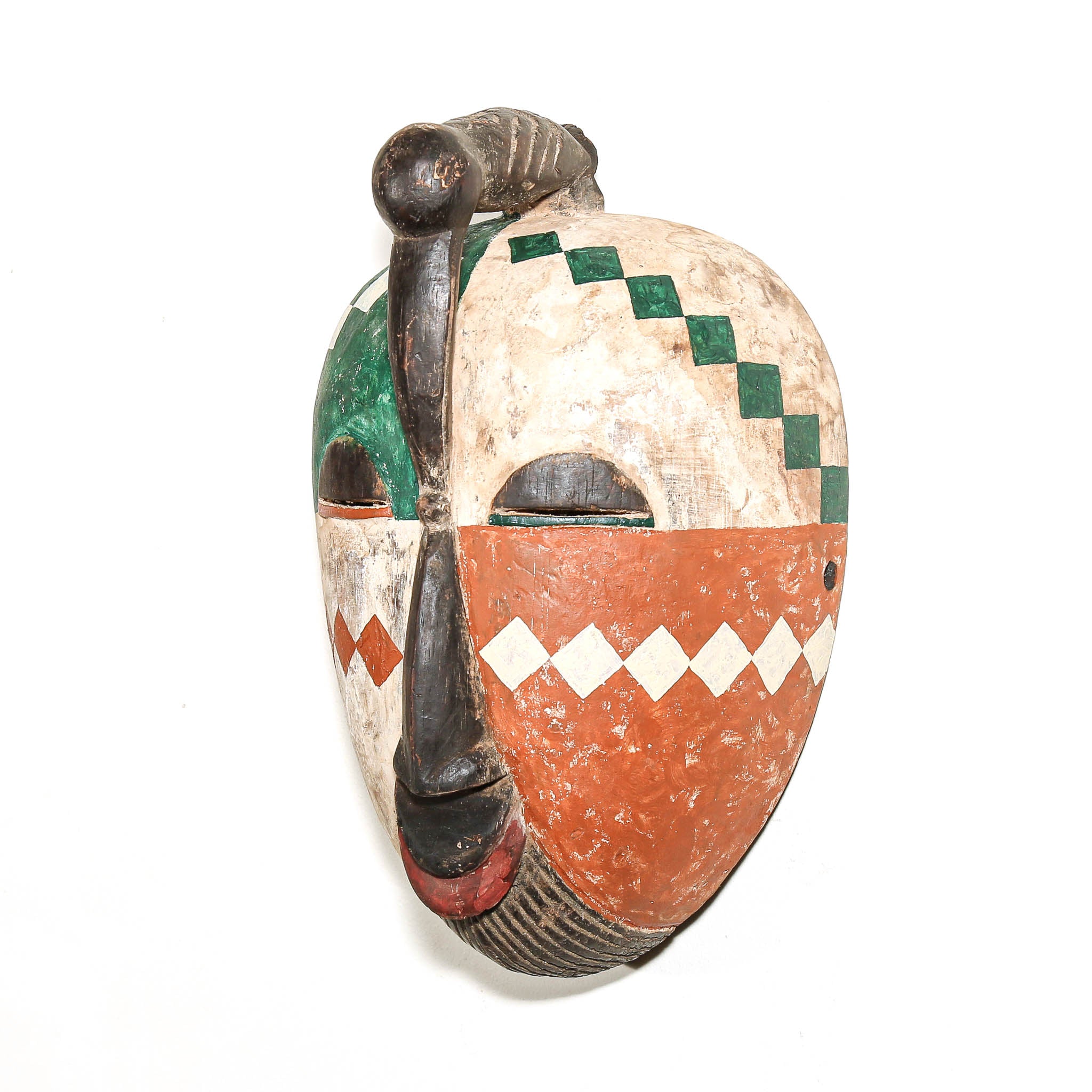 A hand-carved African wooden mask with a prominent vertical nose bar, featuring a cream-colored face with green diamond patterns, a terracotta orange lower section with white diamond accents, and a dark brown antler-like ornament on top.