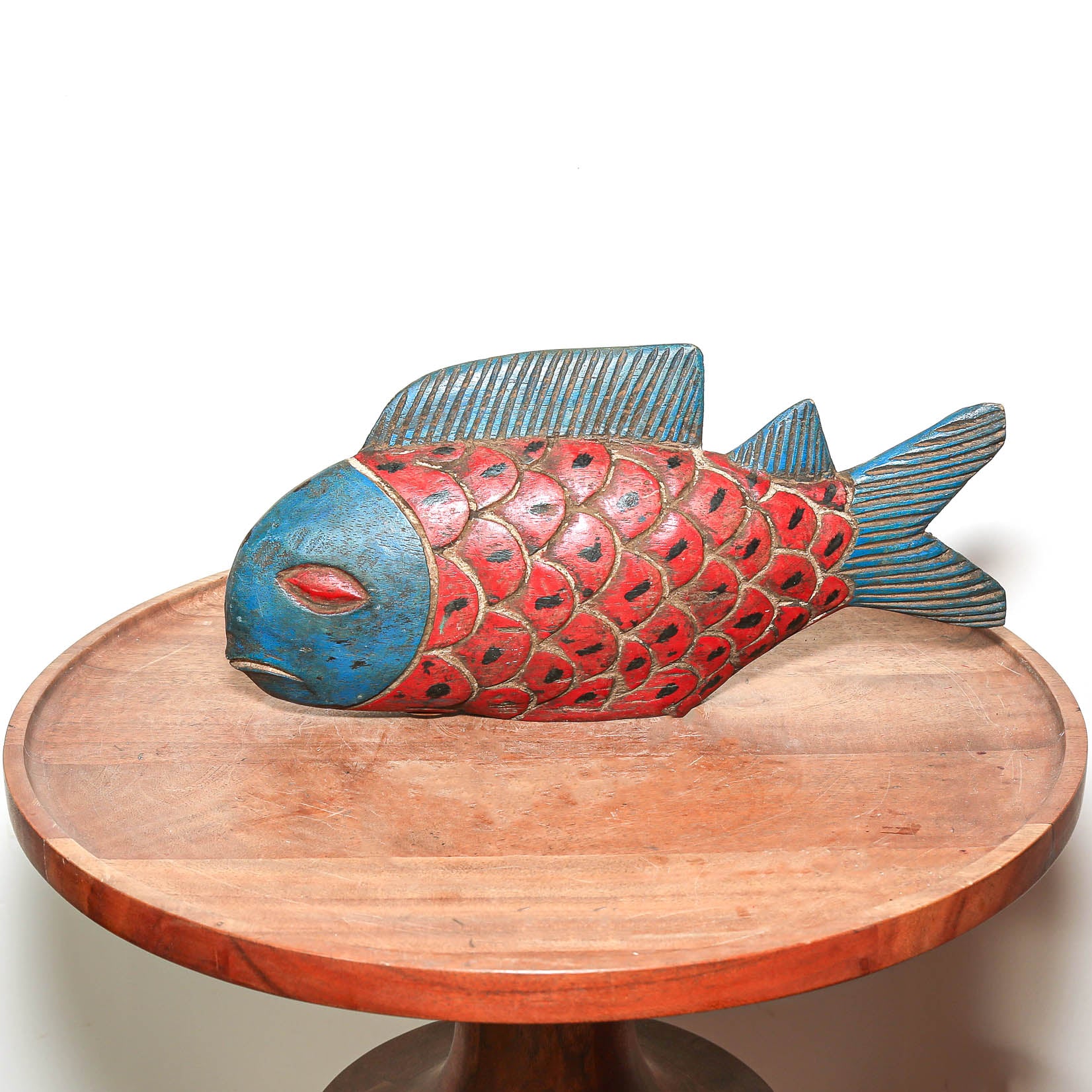 A hand-carved wooden fish sculpture with a red and blue painted finish, displayed on a round wooden pedestal against a white background.