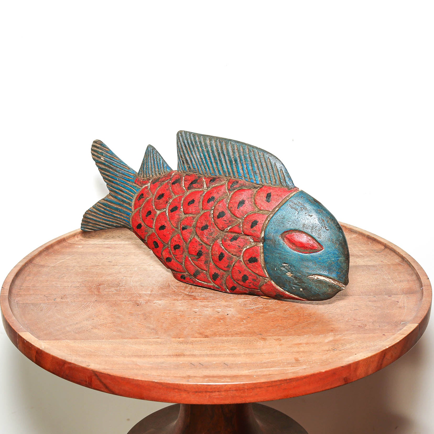 A hand-carved wooden fish sculpture with a red body, blue fins, and black scale details, displayed on a round wooden pedestal against a white background.