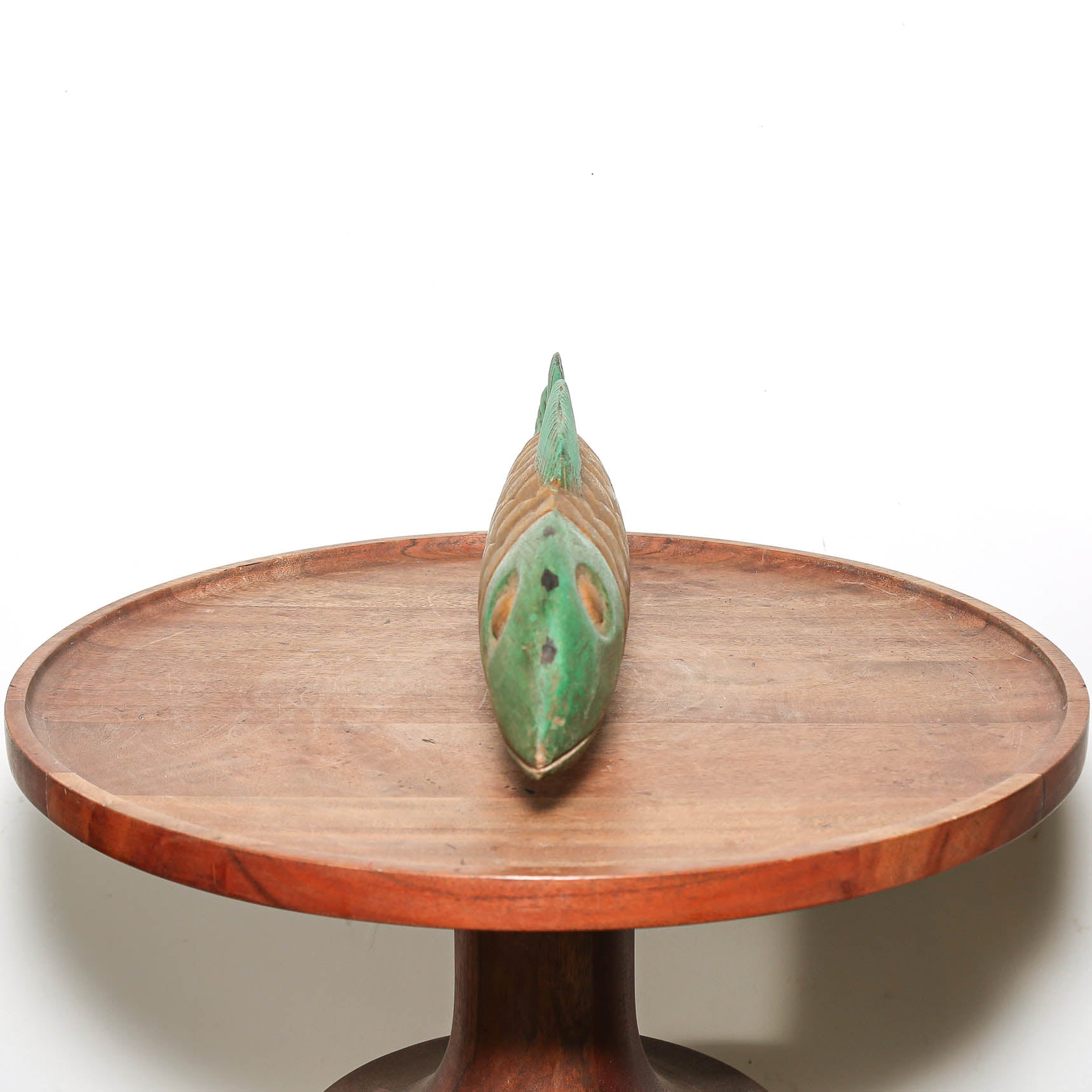 A carved wooden fish-shaped decorative object with a green glazed finish and brown details, displayed on a round wooden pedestal table against a plain white background.