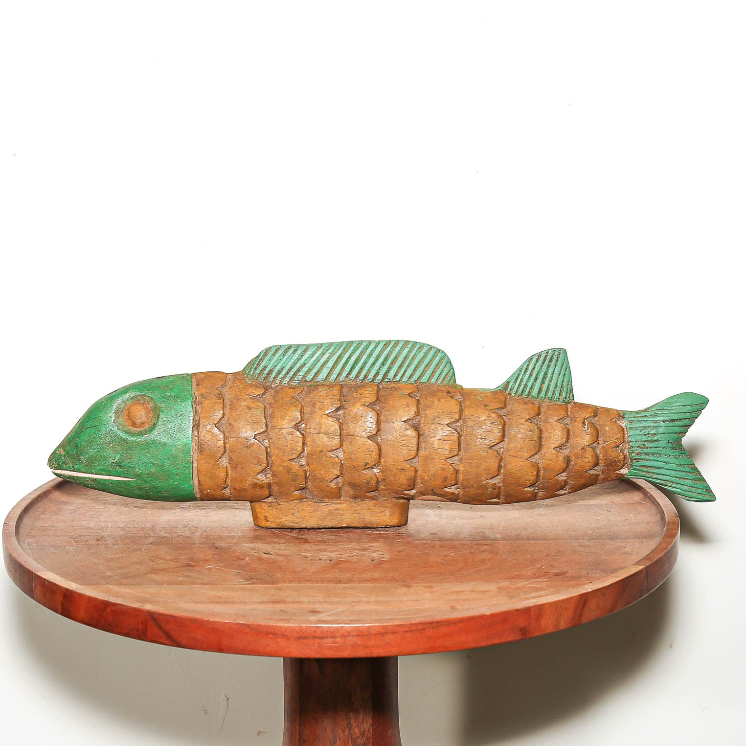 A carved wooden fish sculpture with a green head and tail and a brown scaled body, displayed on a round wooden table against a white background.