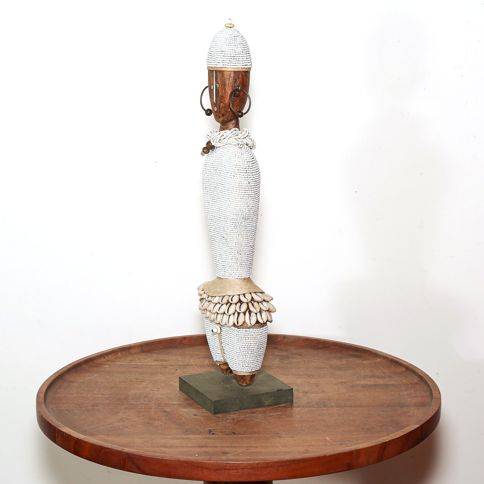 A tall African-style woven figurine sculpture with a white textured body, brown wooden head and feet, and a dark rectangular base, displayed on a round wooden table against a plain white wall.