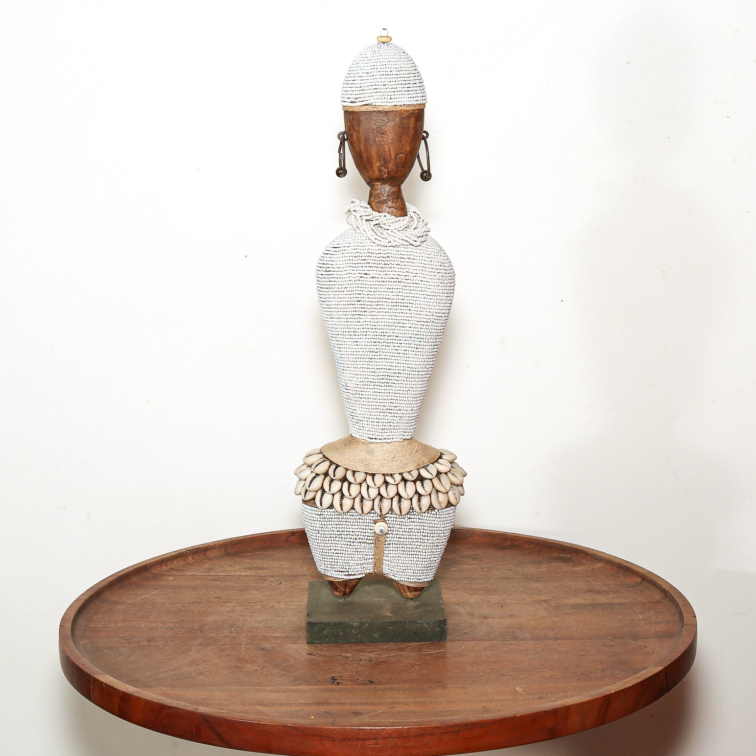 A traditional African wooden doll sculpture with a white beaded body, shell skirt, and wooden head, displayed on a round wooden tray against a white background.