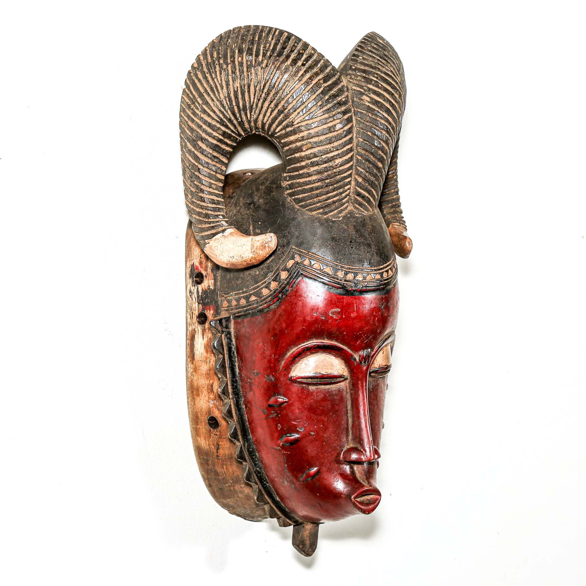 Hand-made African mask with red face and decorative horns on a white wall