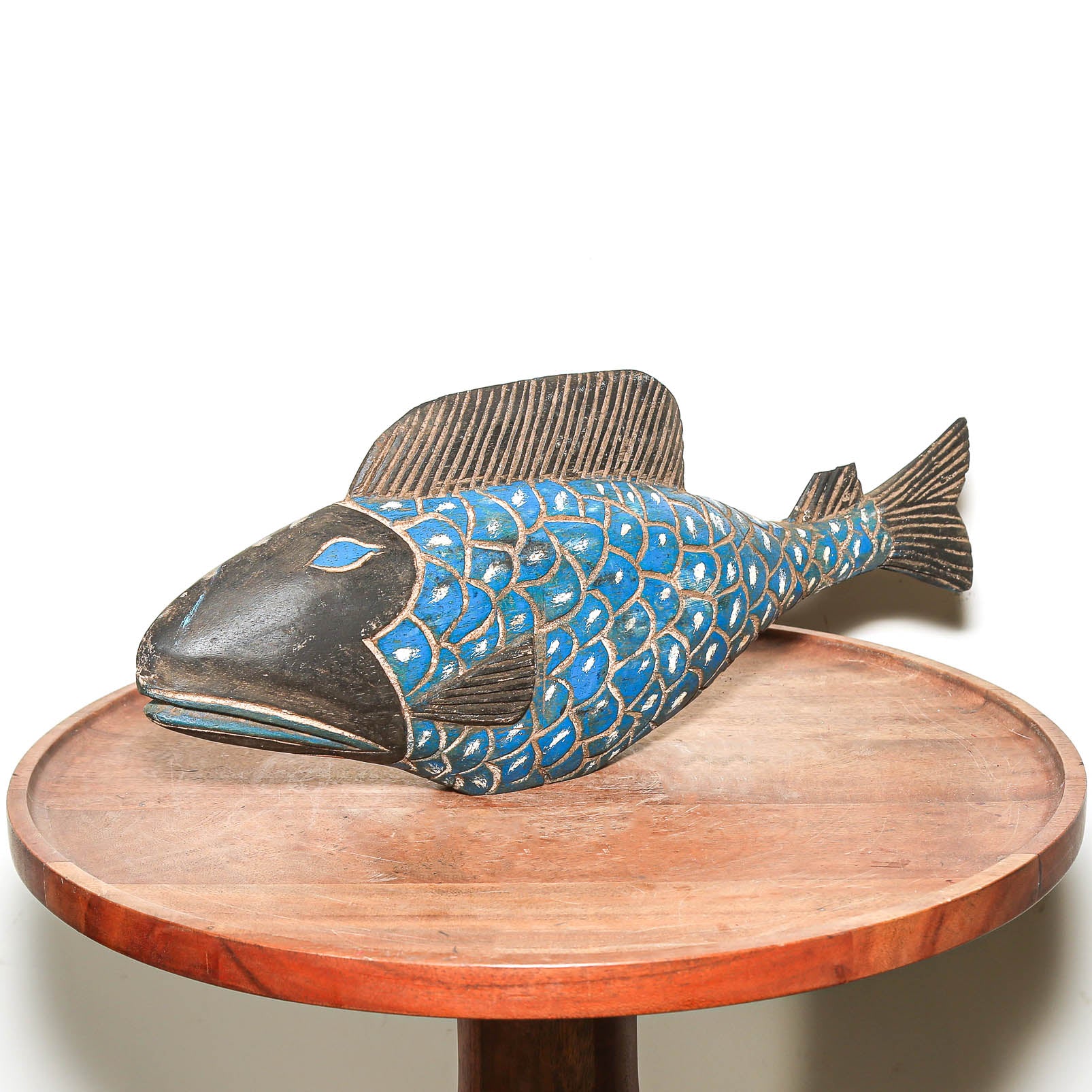 A carved wooden fish sculpture with blue and white painted scales, displayed on a round wooden table against a white background.