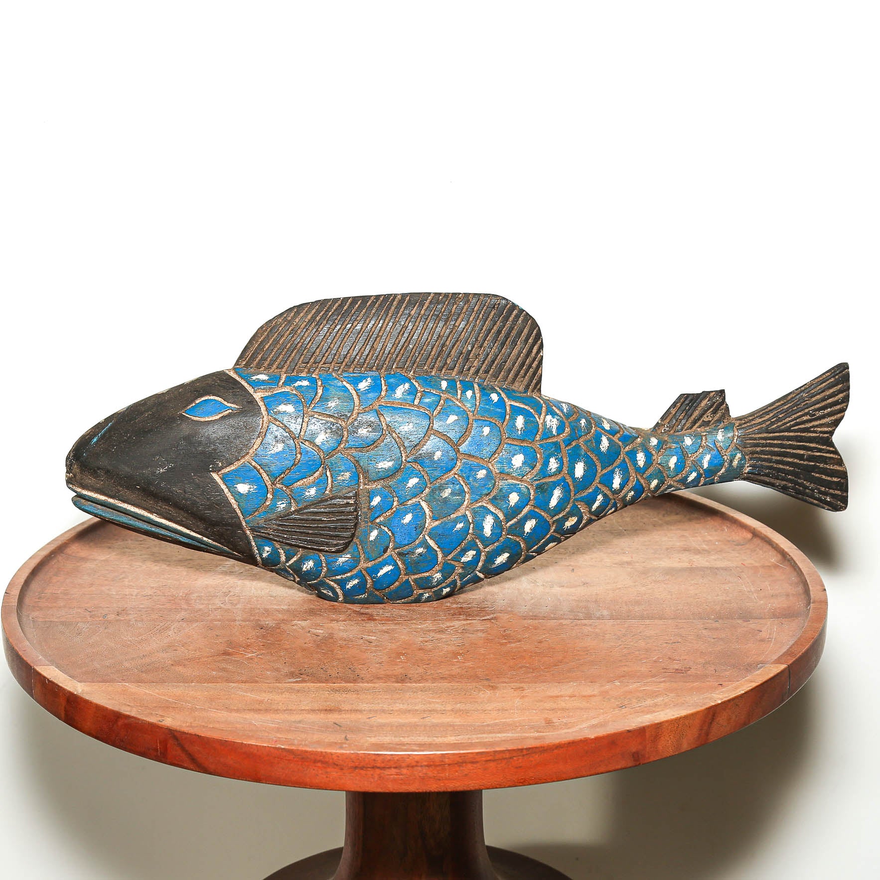 A decorative carved fish sculpture with a blue and black patterned body and dark brown fins, displayed on a round wooden pedestal against a plain white background.