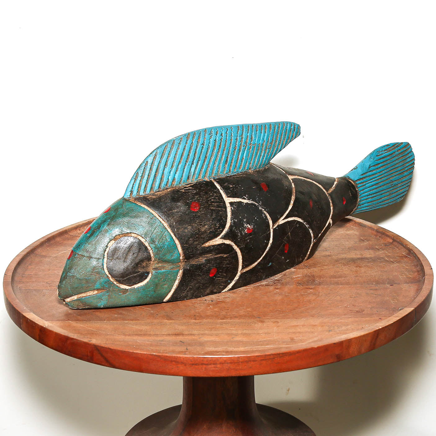 A hand-carved wooden fish sculpture with turquoise blue, black, and red painted details, displayed on a round wooden pedestal against a white background.