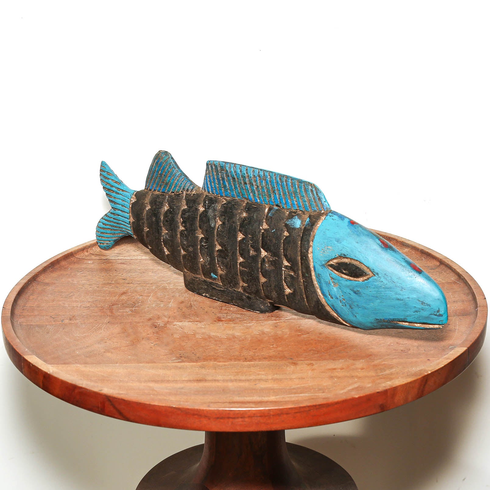 A carved wooden fish sculpture with a bright turquoise blue head and fins, and a dark brown textured body, displayed on a round wooden pedestal against a white background.