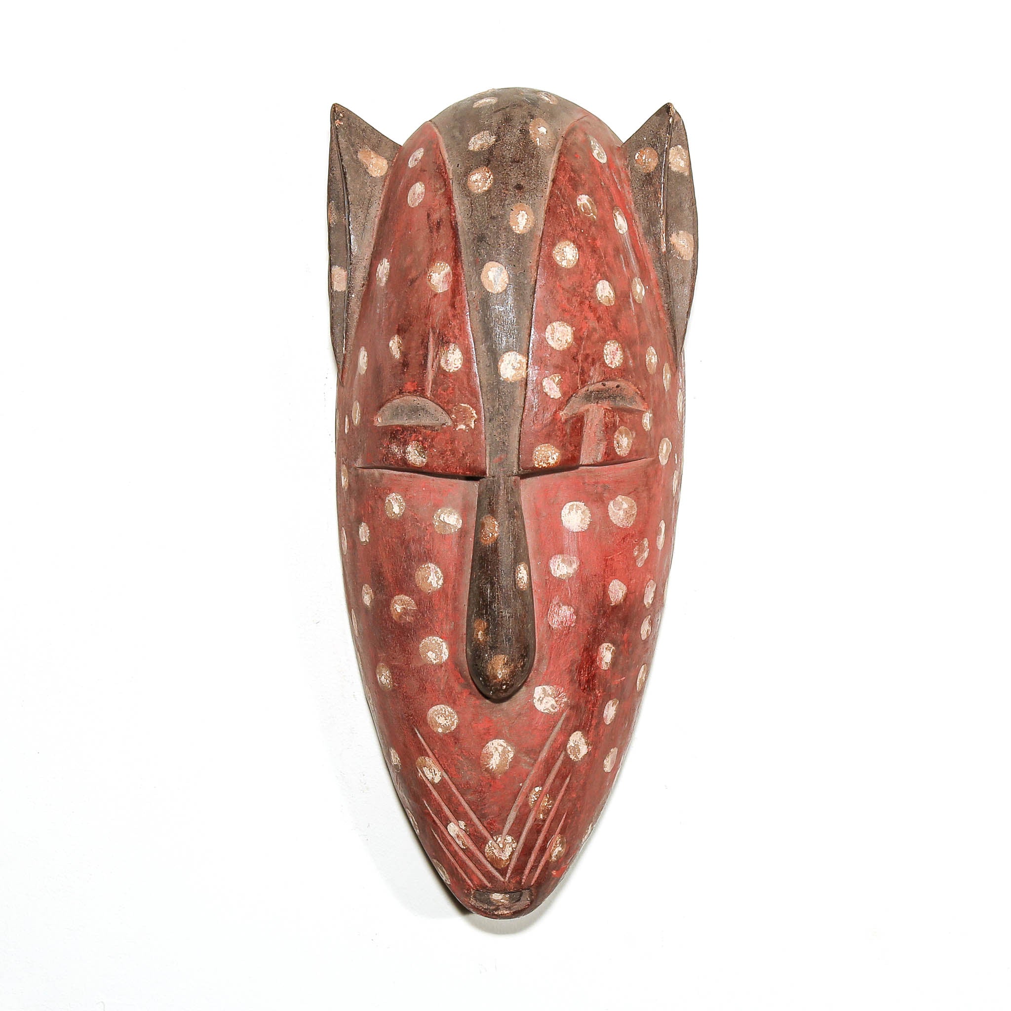 A carved wooden mask with a reddish-brown base, decorated with white speckled dots and a dark brown central vertical feature, photographed against a plain white background.