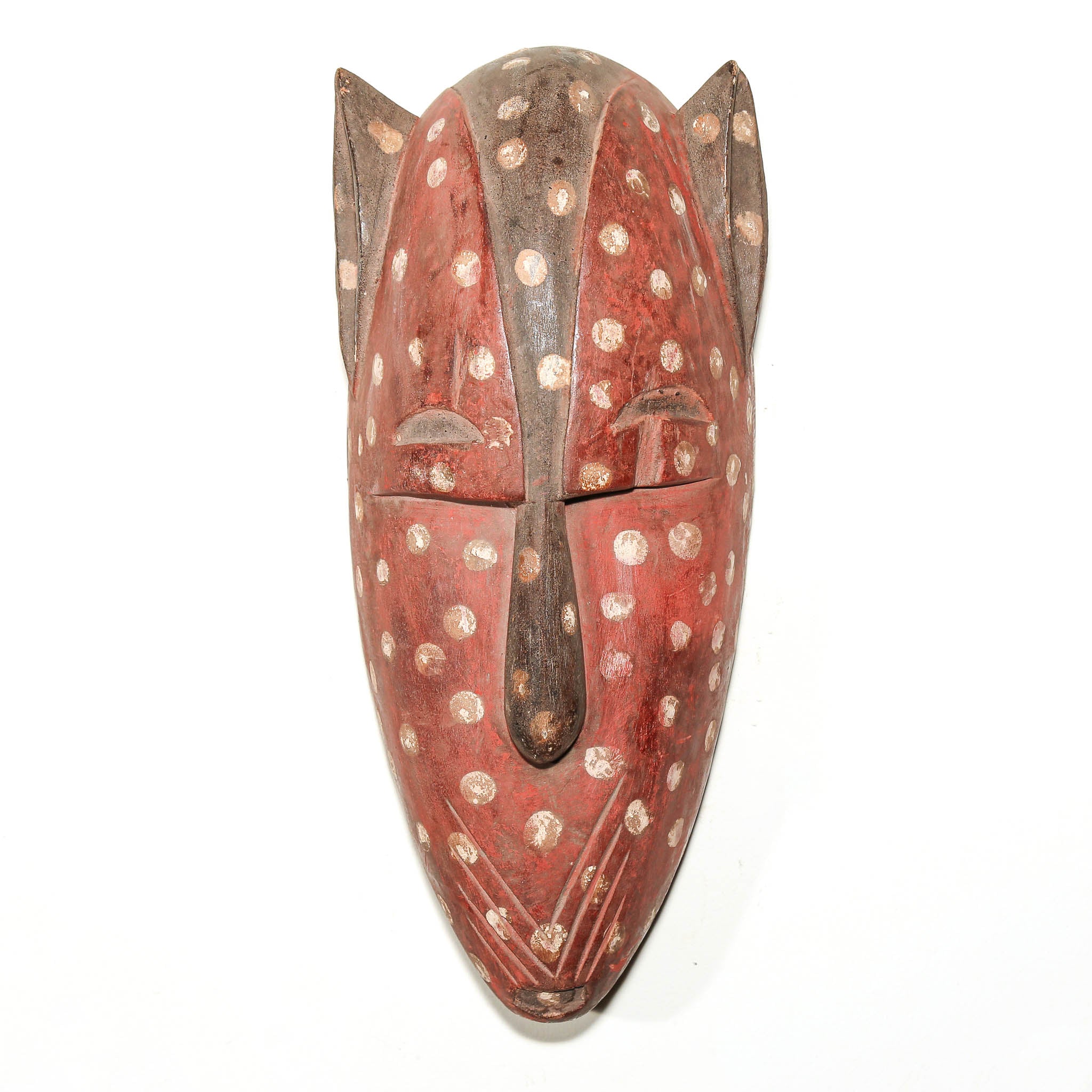 A hand-carved wooden mask with a reddish-brown body, decorated with white and beige circular spots, set against a plain white background.