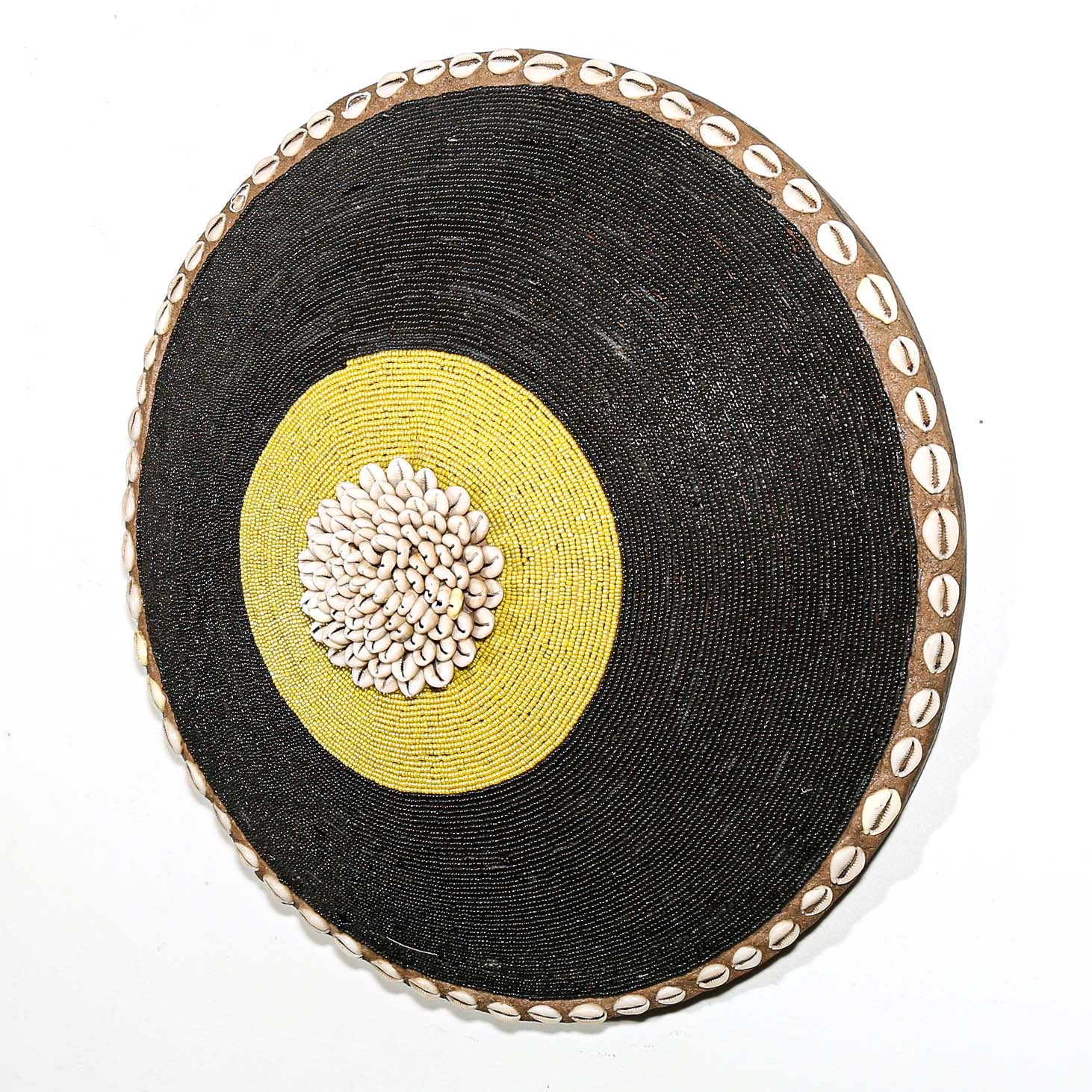 Round woven beaded shield with black surface, yellow center, and decorative edge hanging on a white wall