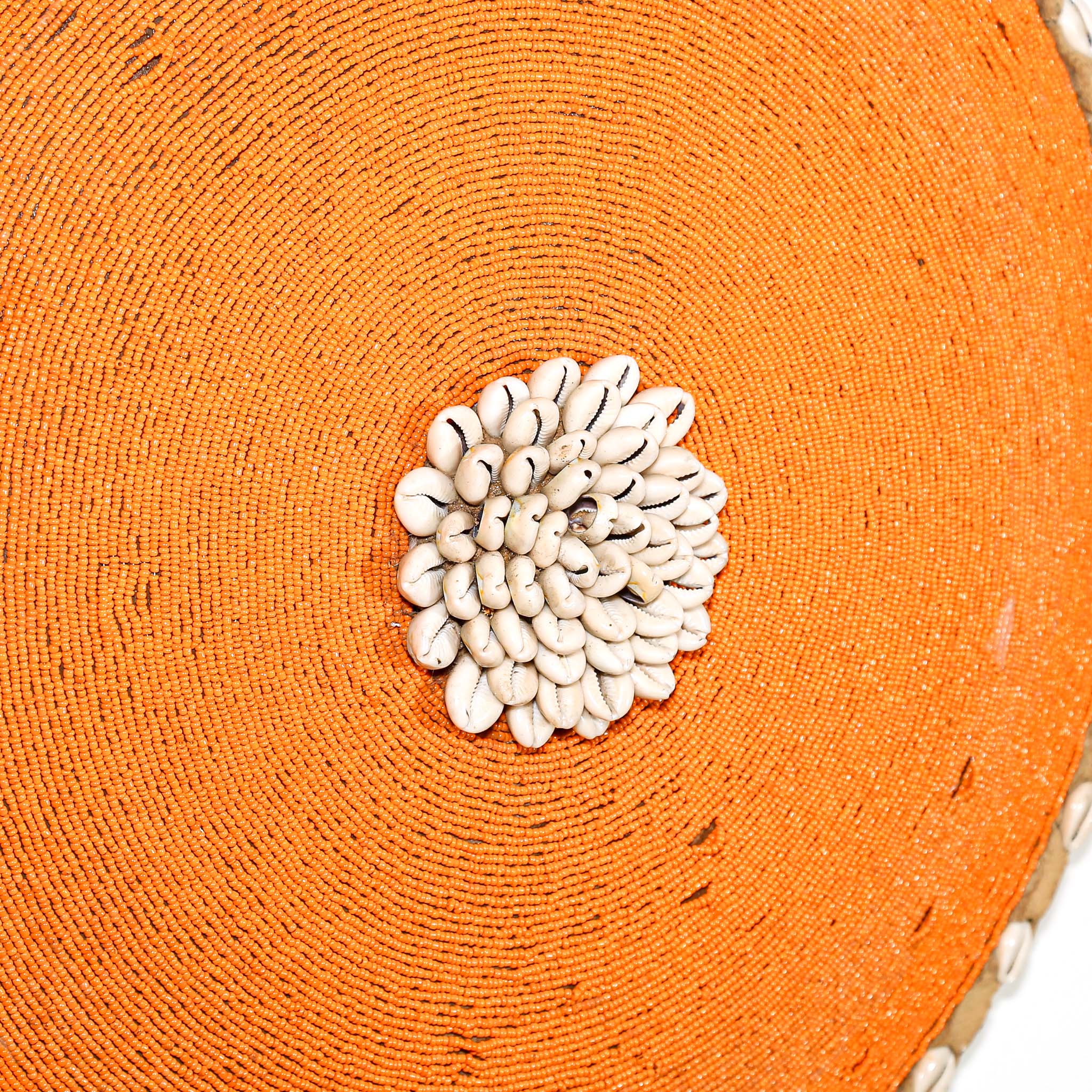 Round orange beaded wall shield decor with a central white flower design made from cowrie shells hanging on a white wall.