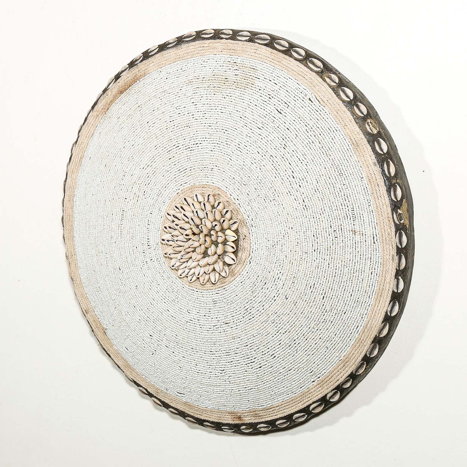 Round white beaded wall shield decor with a central white flower design made from cowrie shells hanging on a white wall.