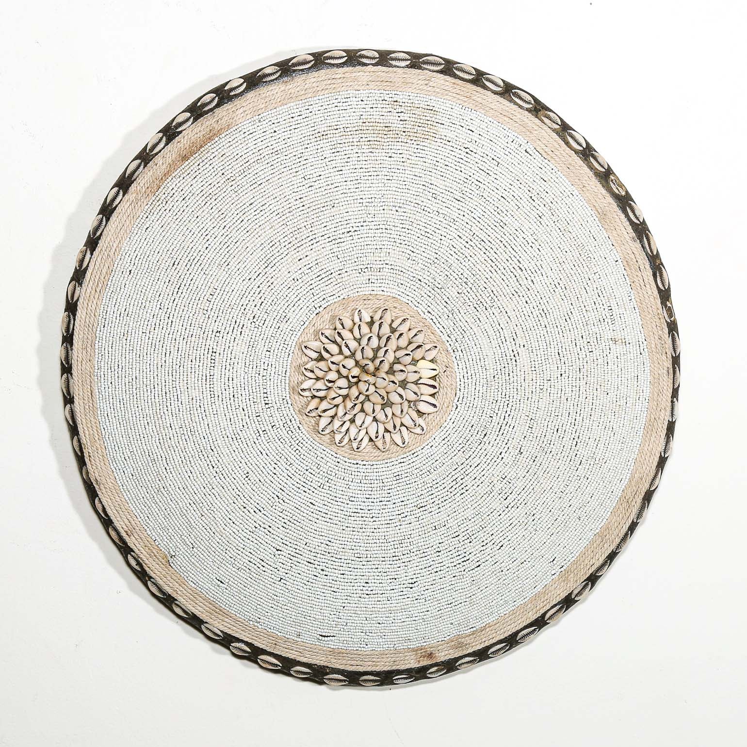 Round white beaded wall shield decor with a central white flower design made from cowrie shells hanging on a white wall.