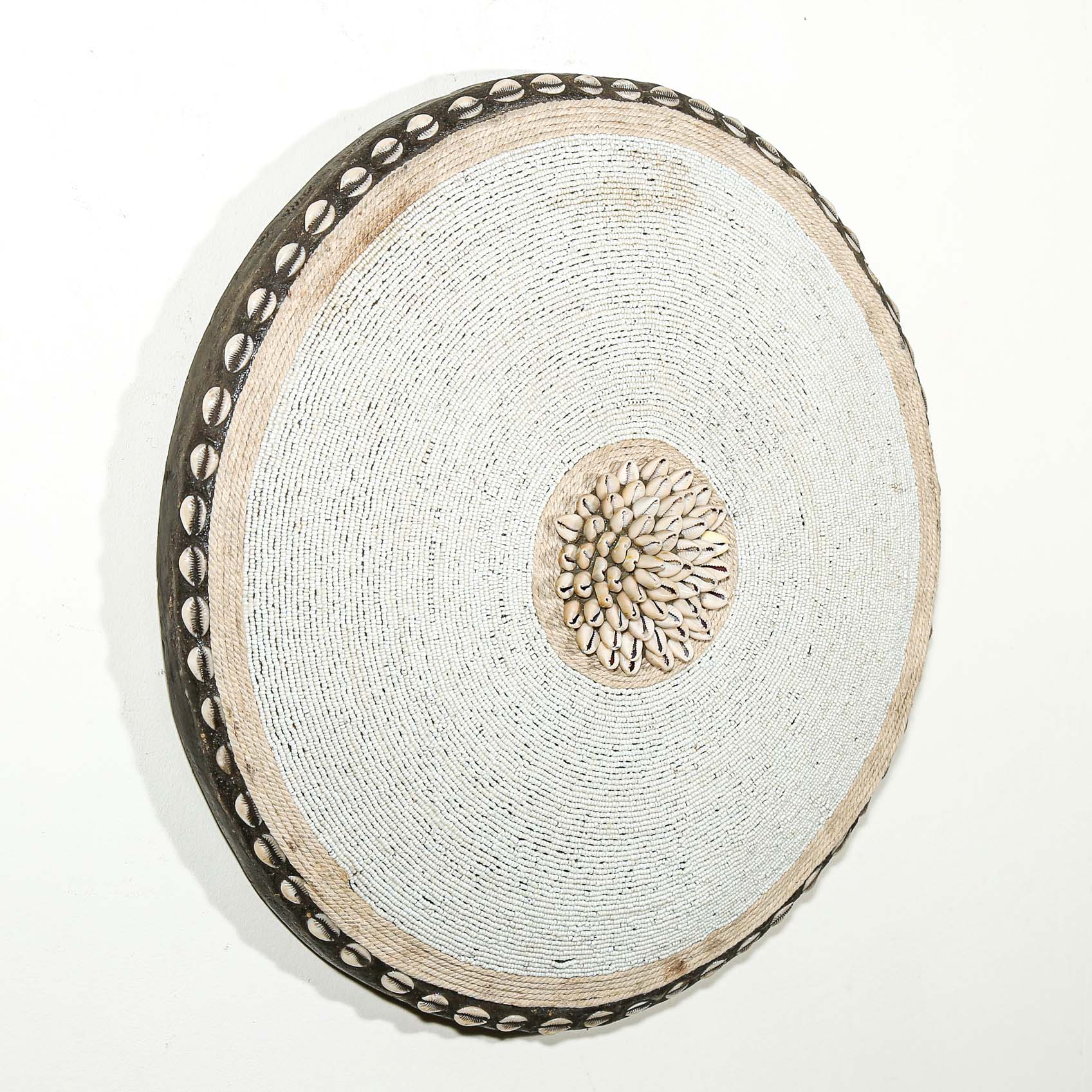 Round white beaded wall shield decor with a central white flower design made from cowrie shells hanging on a white wall.