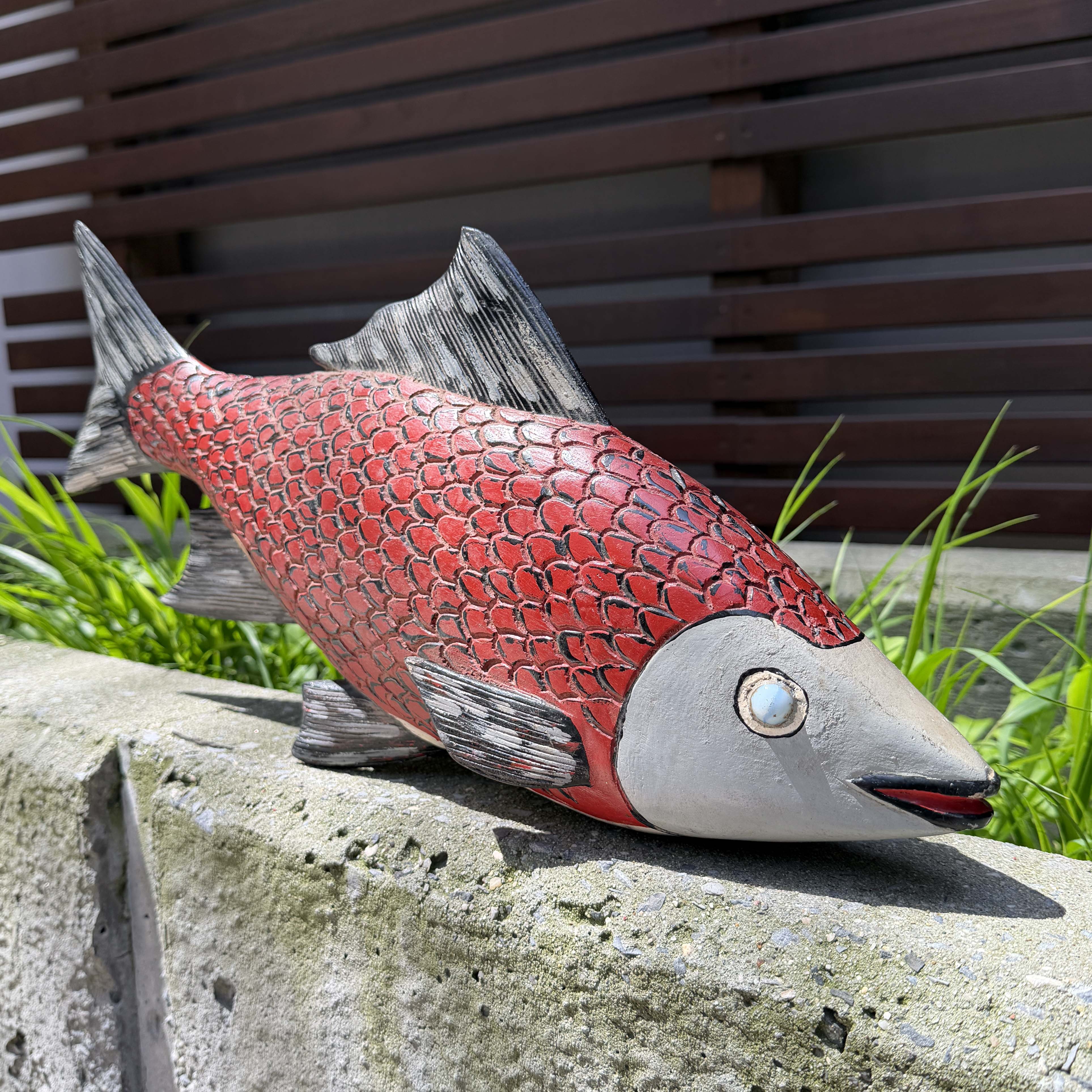 Red and gray fish sculpture on a stone ledge with grass and wooden background