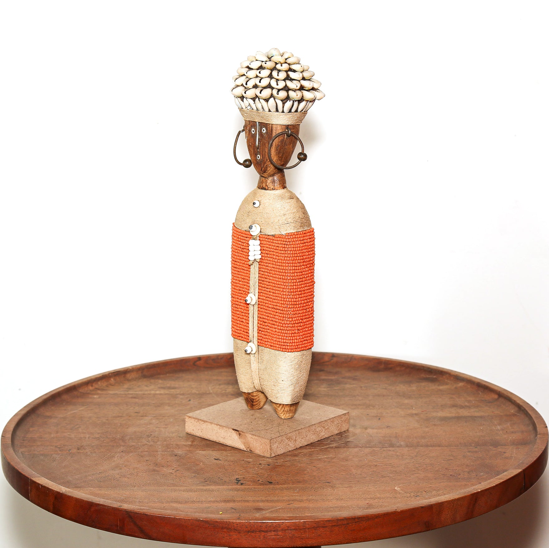 A handmade African-style wooden doll figurine with a woven orange and beige body, standing on a small wooden base on a round wooden tray against a white background.