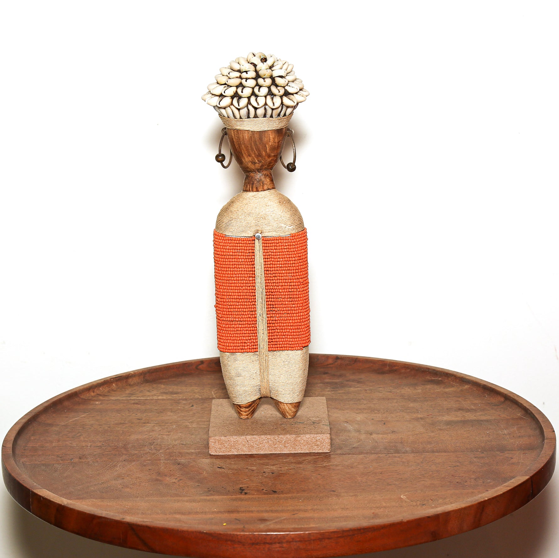 A handcrafted African-style wooden doll sculpture with a woven orange body, standing on a small rectangular base and placed on a round wooden tray against a plain white background.