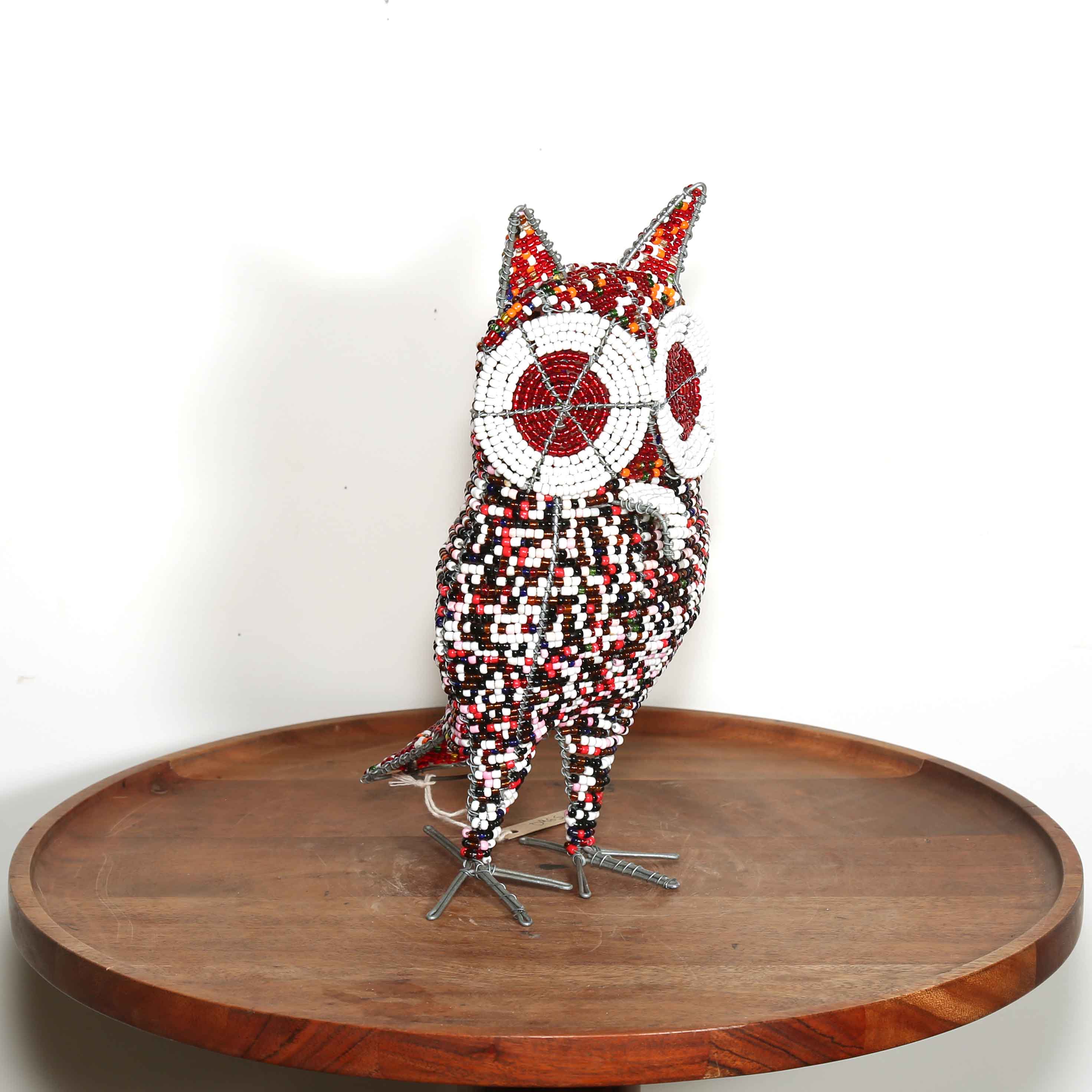 Decorative owl sculpture on a wooden platform with a white background. The owl has red and white eyes made from beads