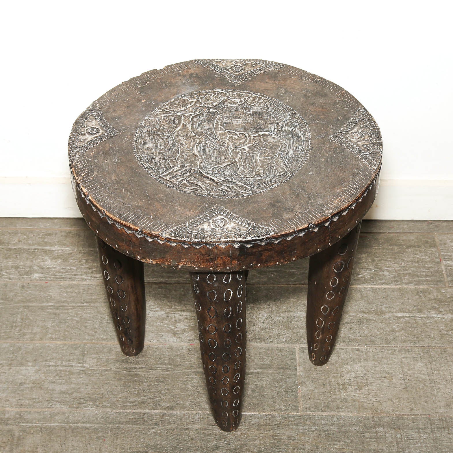 A rustic carved wooden stool with a round seat featuring an embossed animal motif and three tapered legs with circular cutout patterns, set on a gray floor against a white wall.