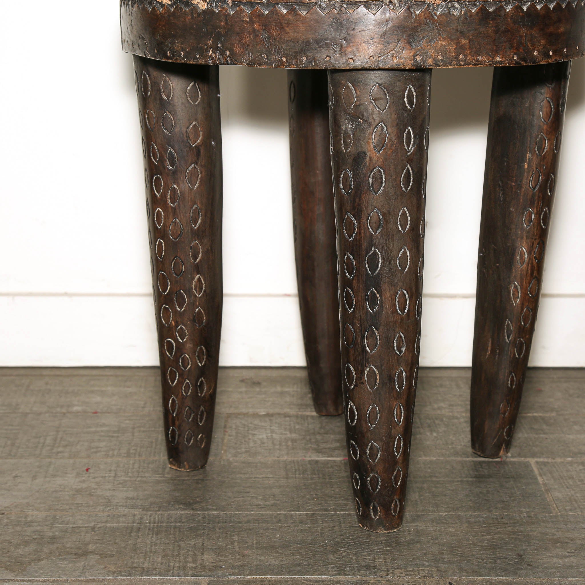 Close-up of a dark carved wooden stool or table leg detail, showing three tapered legs with circular cutout patterns and a rustic finish, set against a white wall and gray floor.