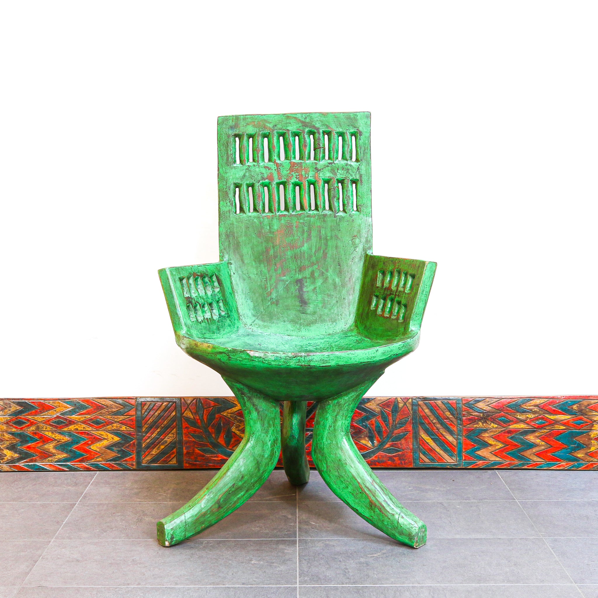 A green wooden lounge chair with a distressed, weathered finish, featuring a high slatted backrest and armrests, set against a white wall with a colorful patterned tile border.