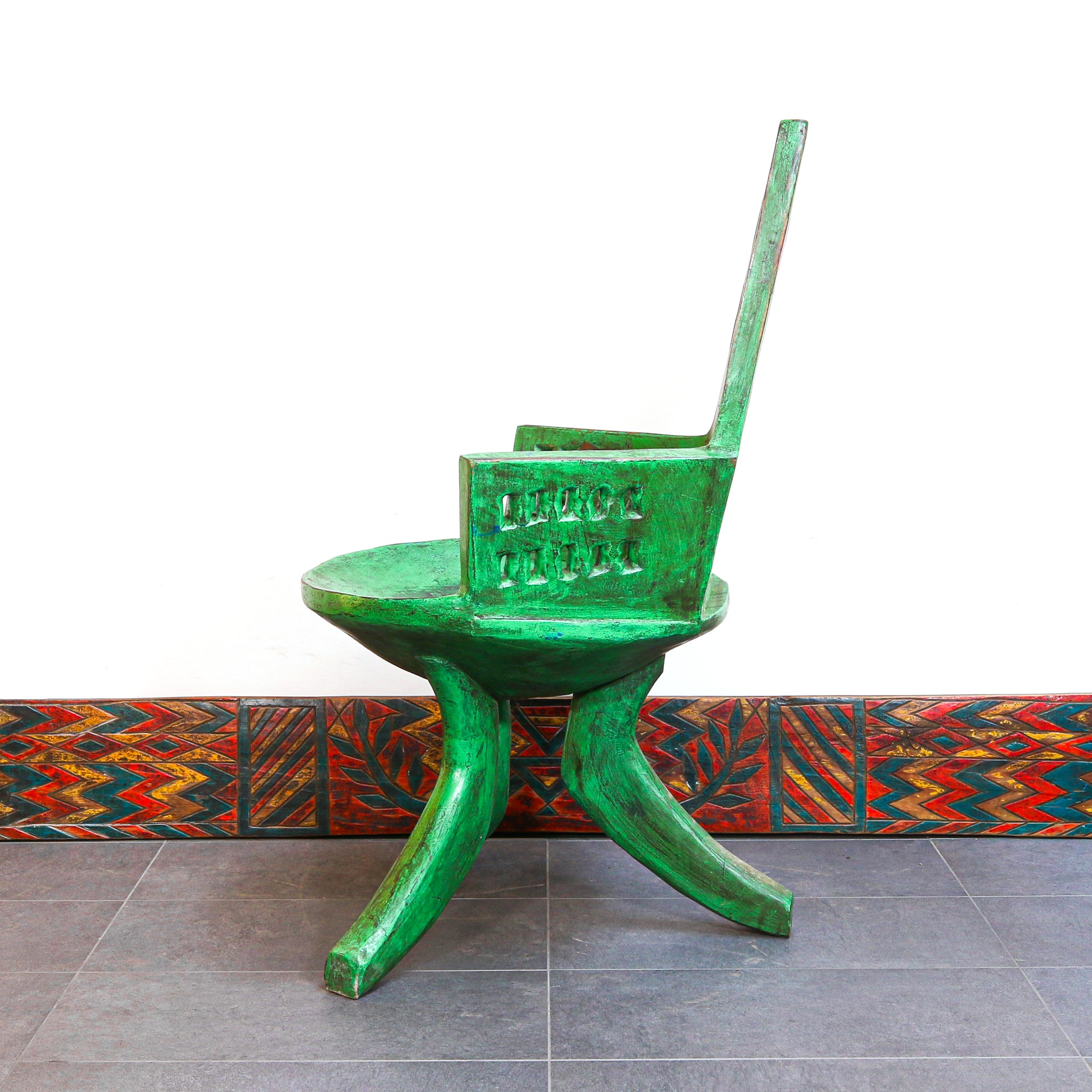 A green sculptural wooden chair with a high back and carved details, set against a white wall with a colorful patterned tile base.