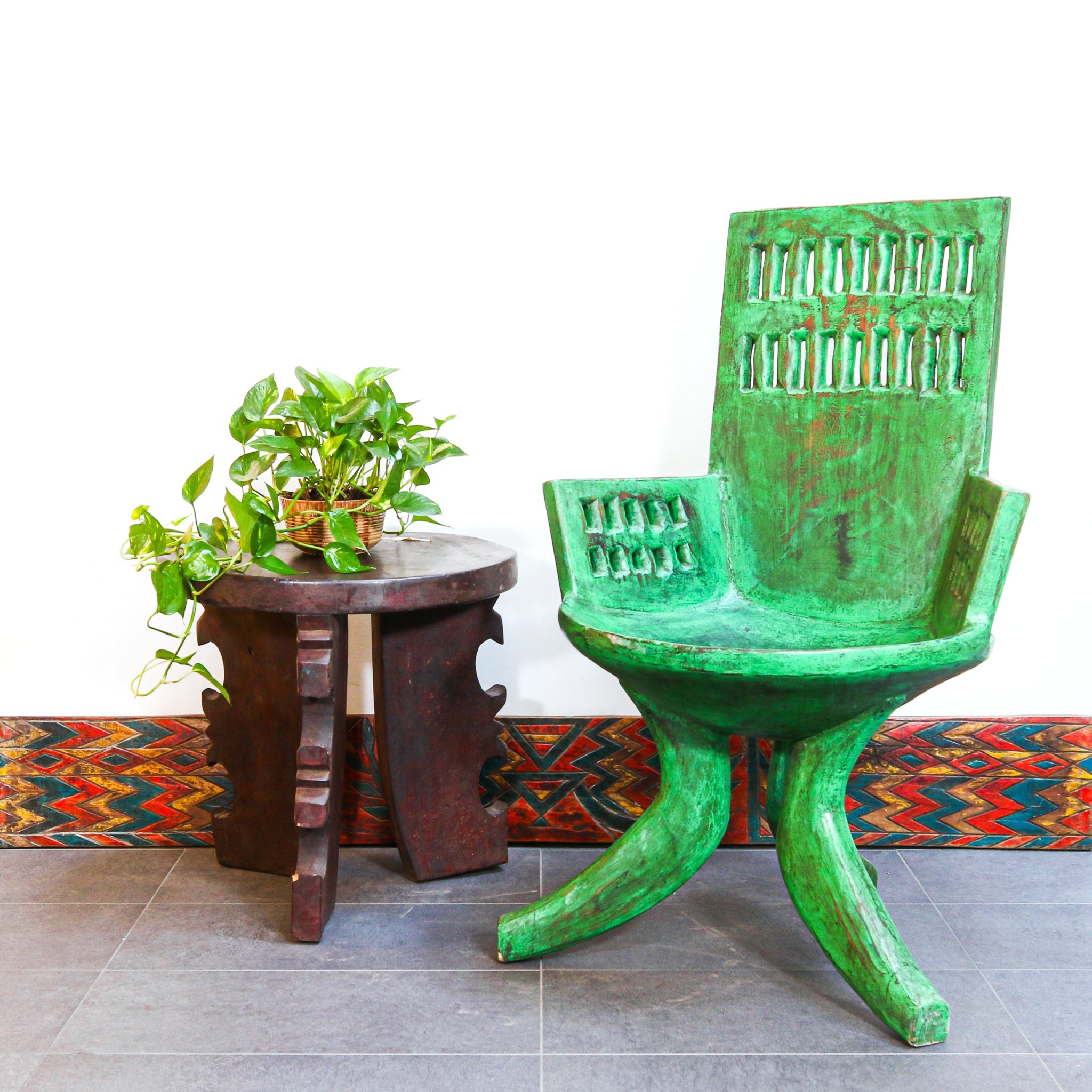 A green distressed wooden armchair with carved details, shown next to a small dark wooden side table holding a potted green plant, set against a white wall with a patterned rug on a gray tiled floor.