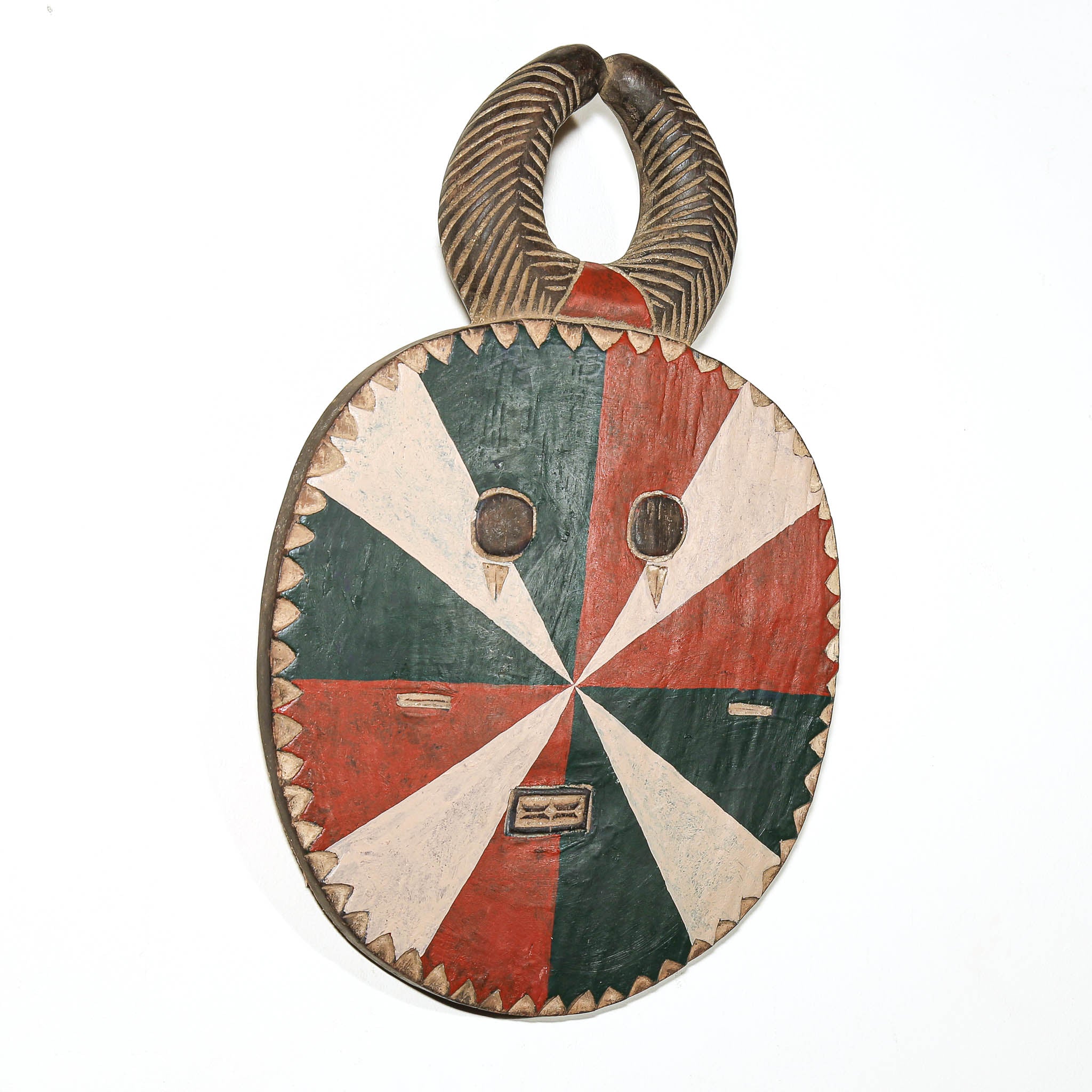 A hand-carved African wooden mask with a curved horn-like handle, featuring a starburst pattern in red, dark green, and beige on the face, with carved facial features and a serrated wooden edge.